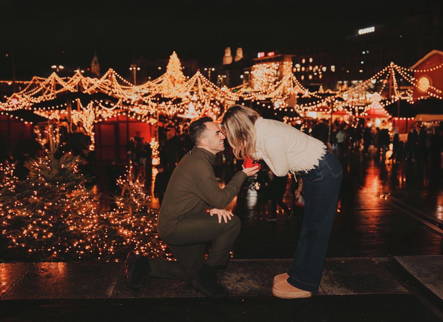 Man kneeling on one knee proposing to woman at glowing Christmas market at night.