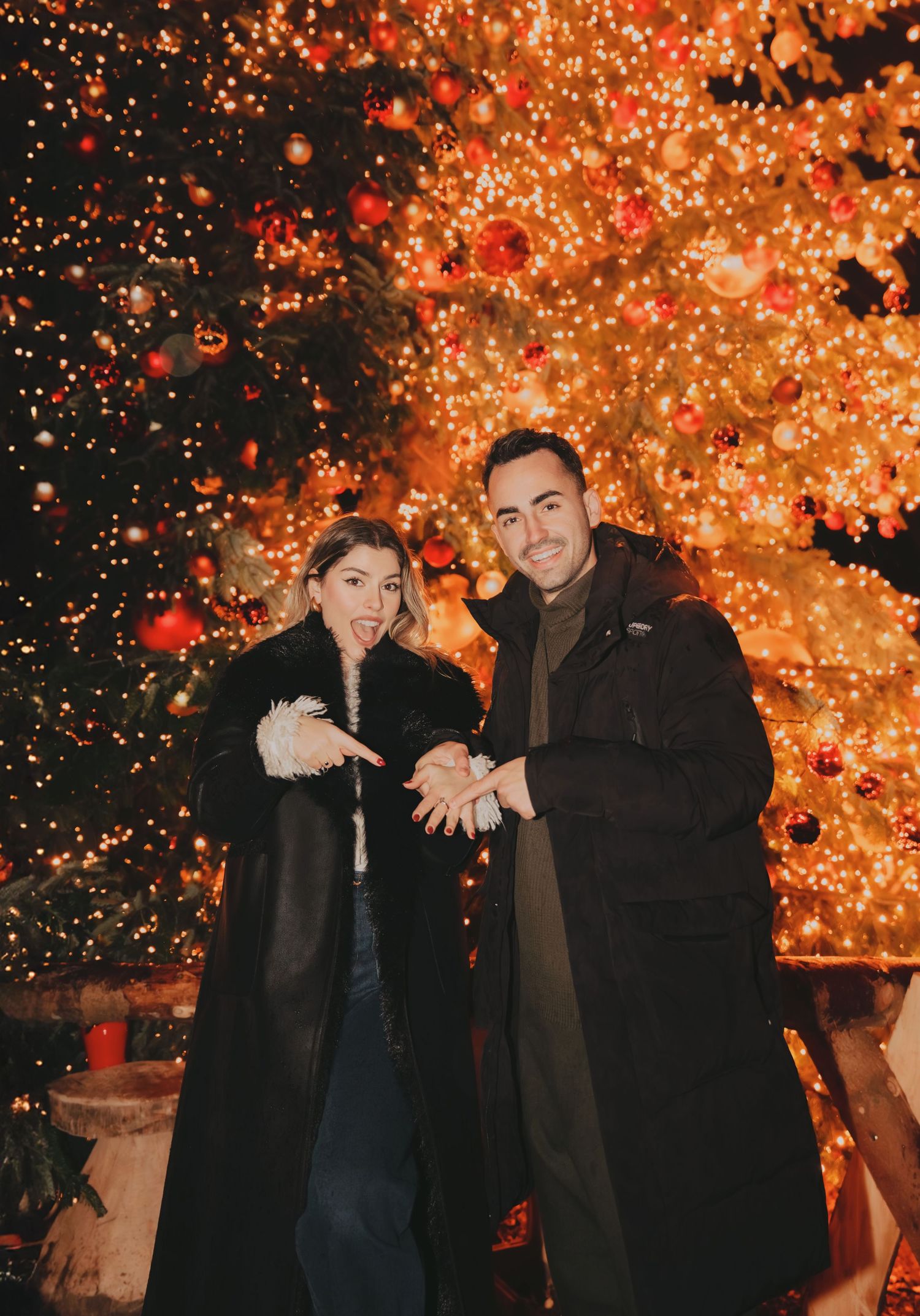 Couple posing in front of glowing Christmas tree with red ornaments at night.