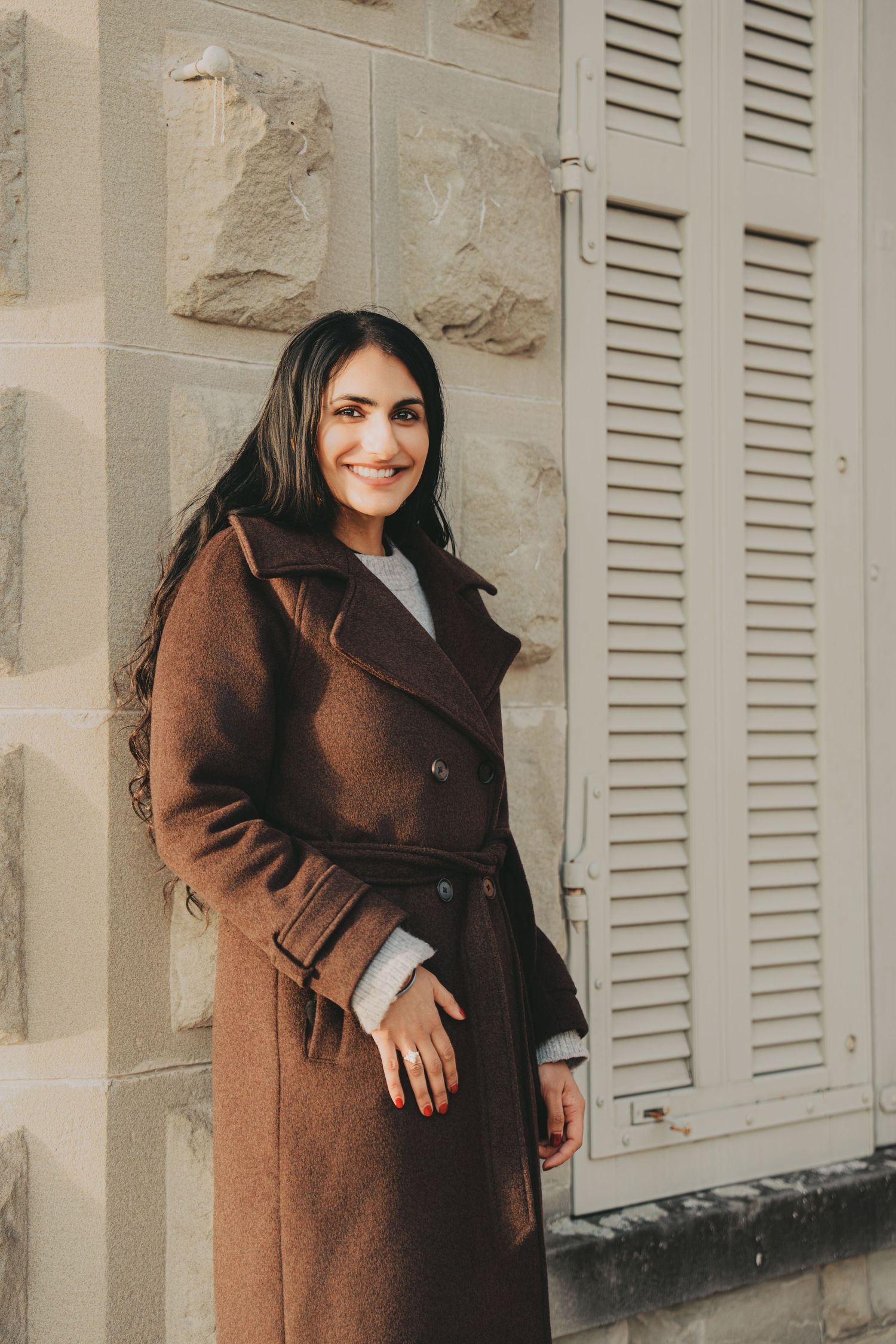 Smiling South Asian woman in brown coat leaning against stone wall beside a shuttered window.