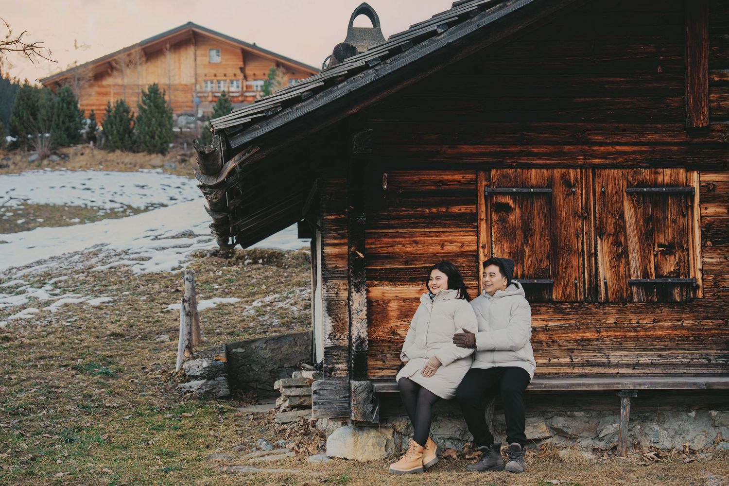 Couple sitting together on bench outside rustic wooden alpine chalet at golden hour.