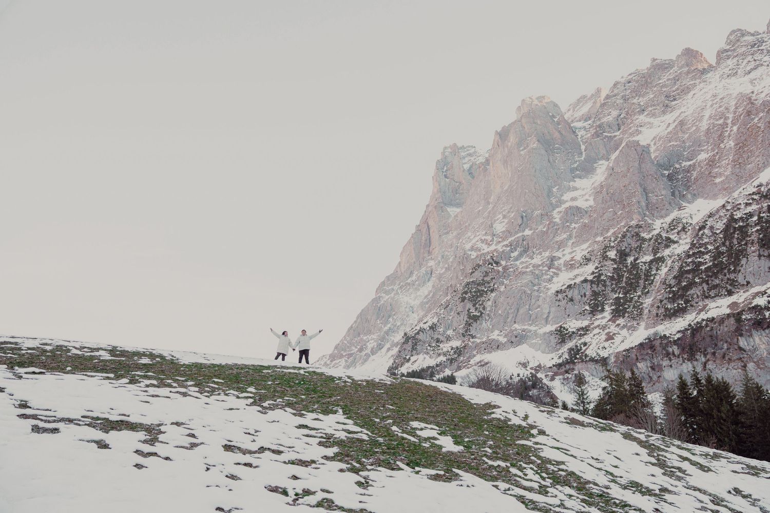 Two people standing on snowy hillside with dramatic rocky alpine mountain in background.