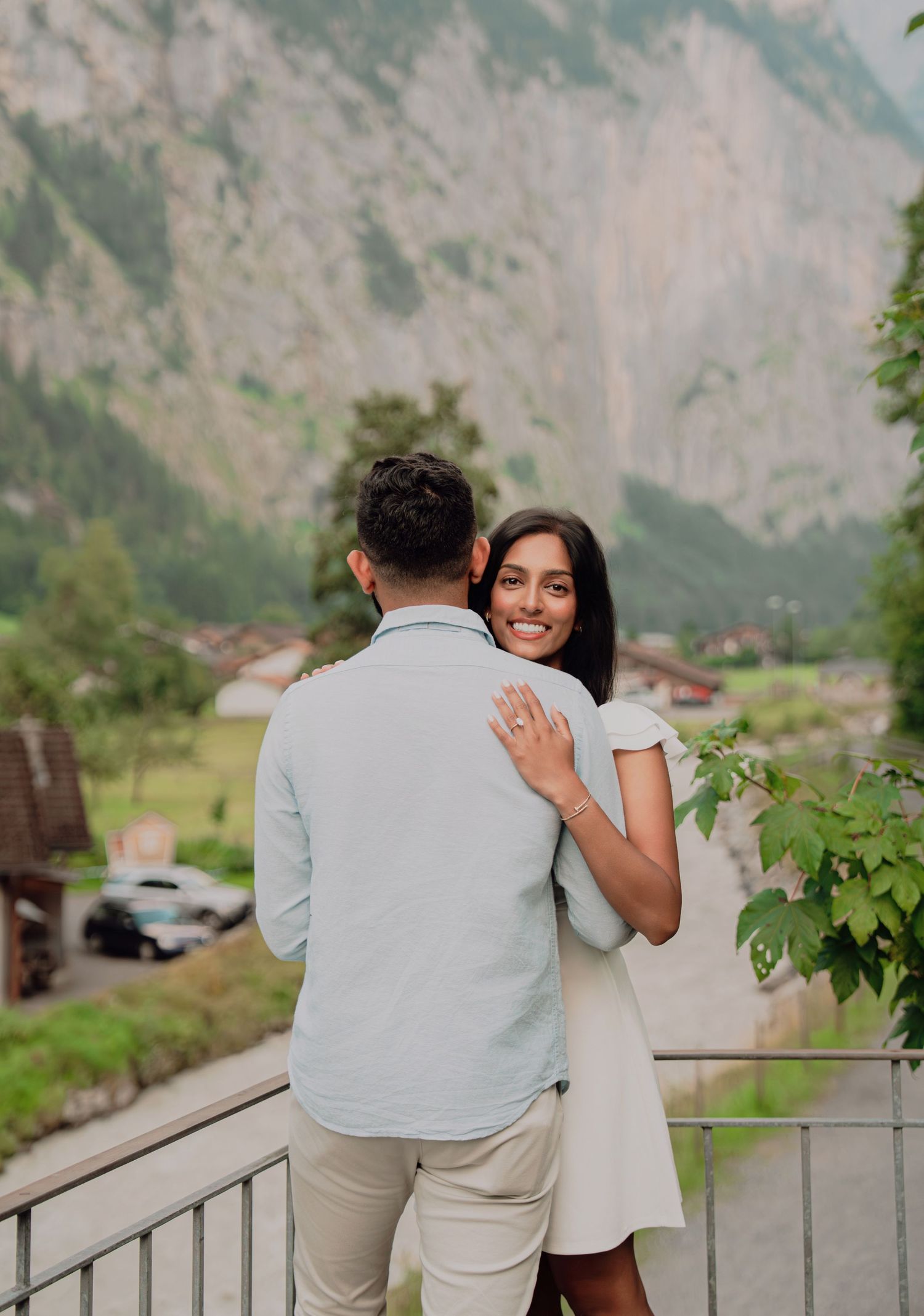 Smiling woman embraces her fiance showing engagement ring with Swiss Alps in the background.