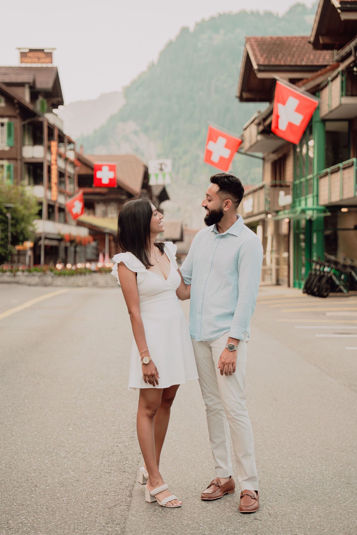 Couple gazing at each other on empty Swiss village street lined with traditional buildings and red Swiss national flags.