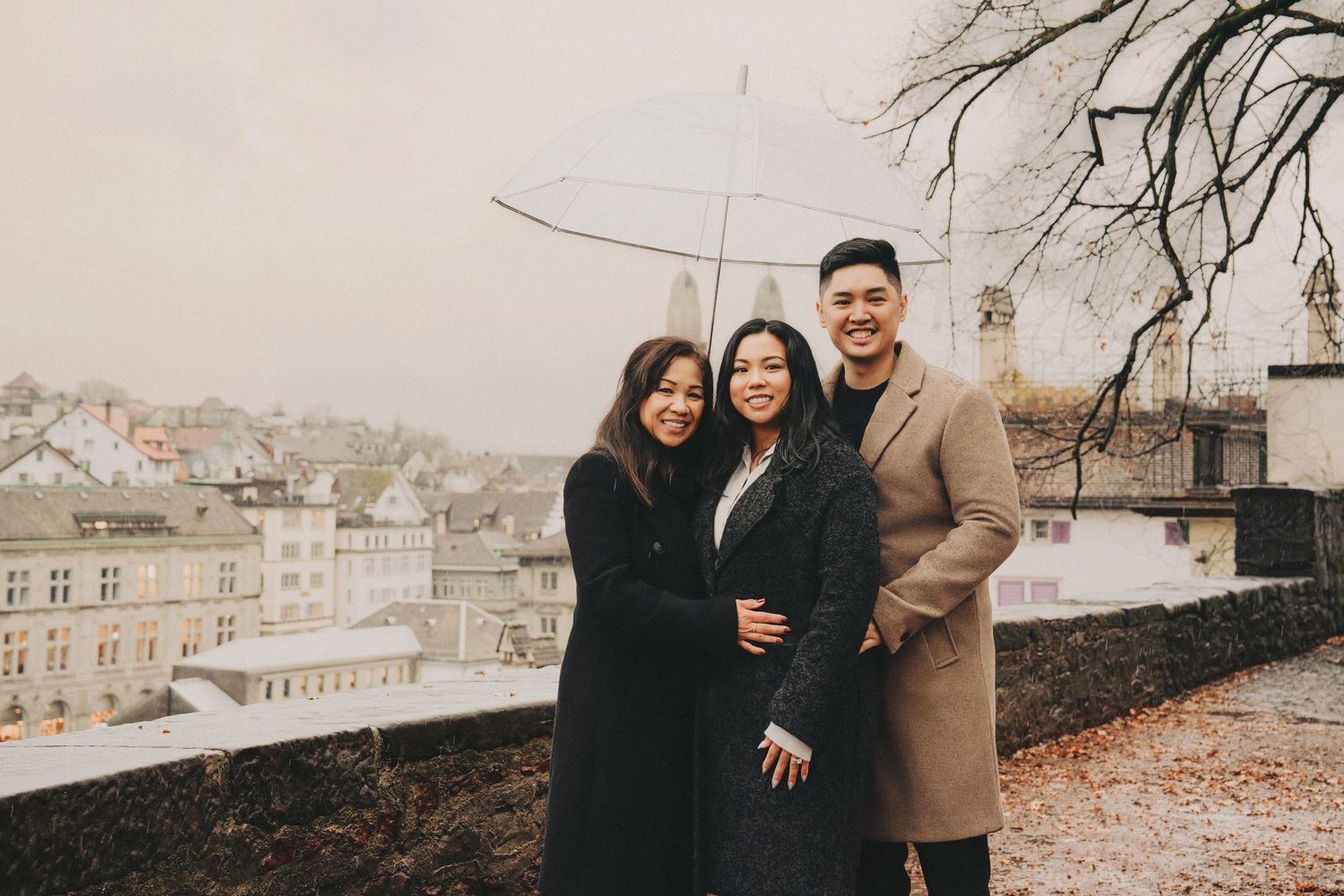 Three friends smile under a clear umbrella on a rainy day with a European cityscape in the background.