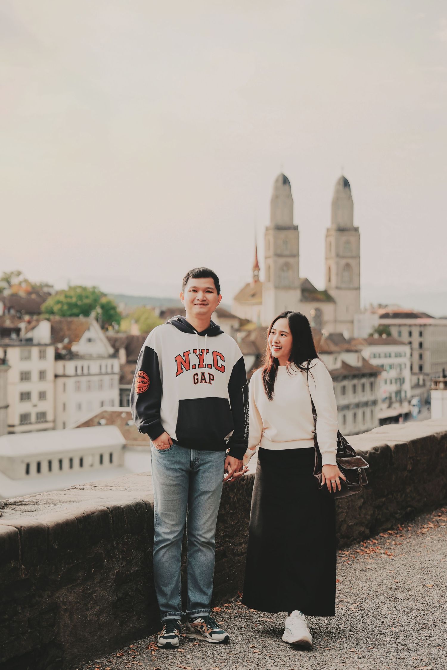 Couple holding hands with Grossmünster cathedral and Zurich skyline backdrop, man in NYC Gap hoodie.