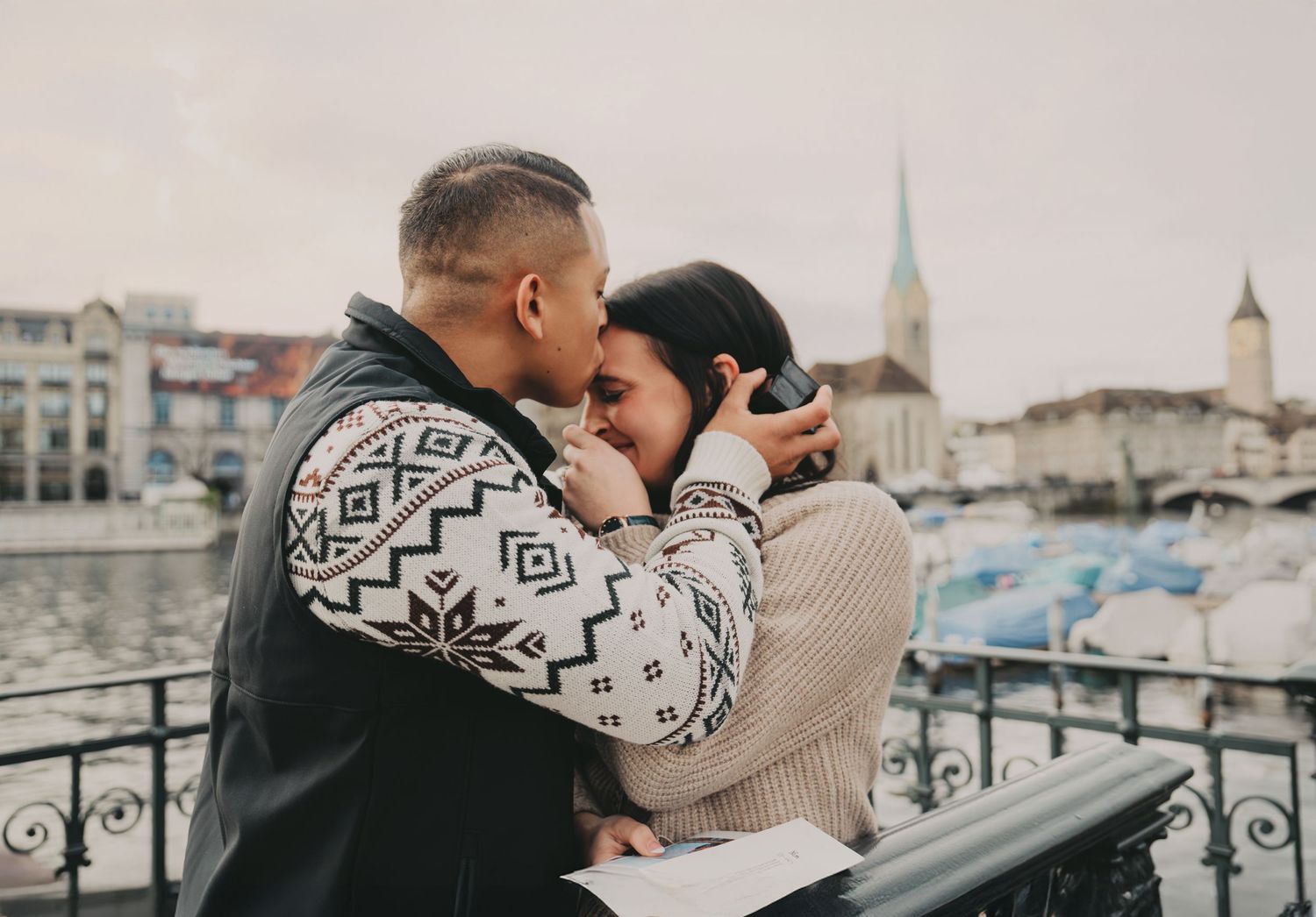 Couple sharing intimate kiss on Zurich waterfront bridge with historic church spire backdrop.