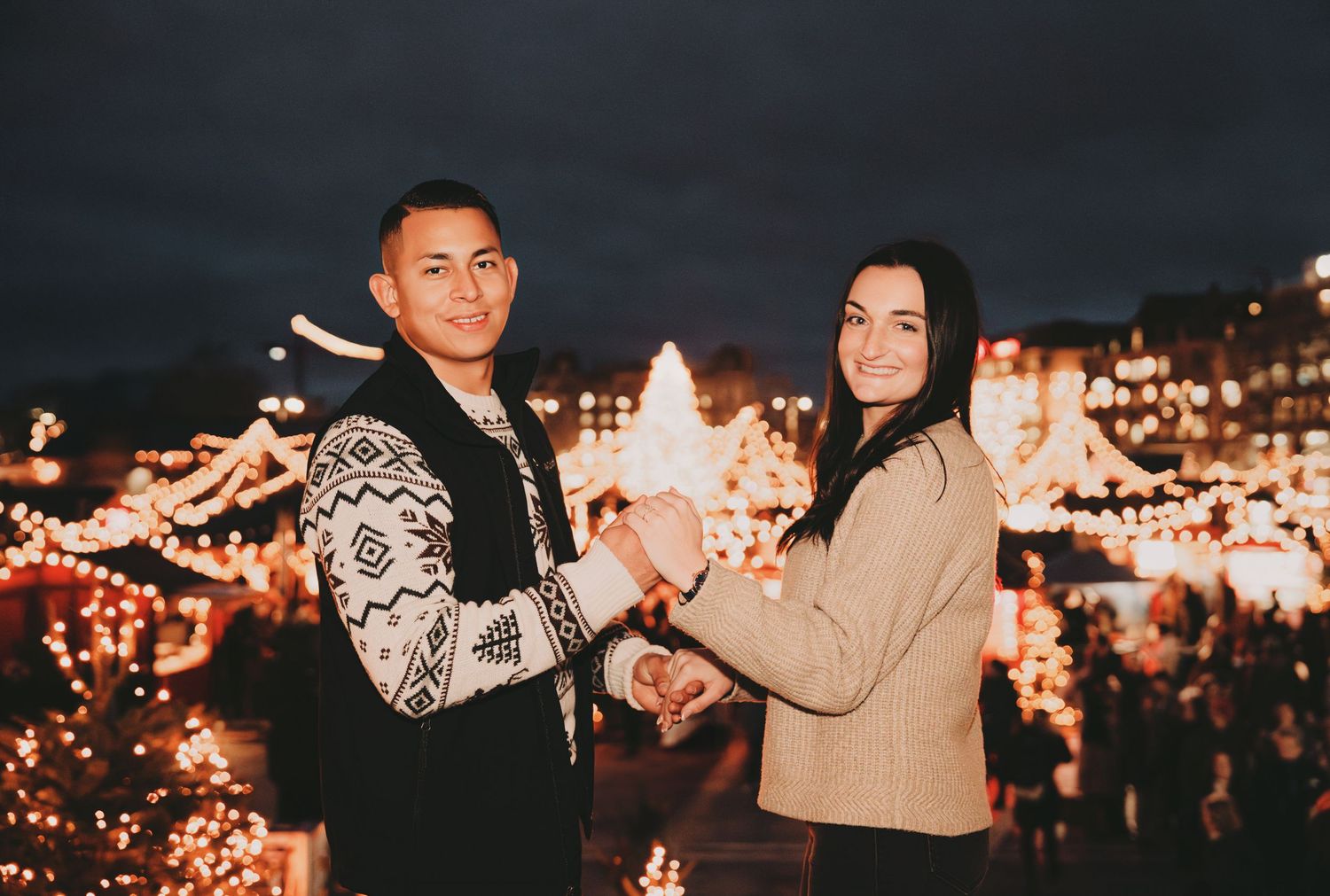 Happy couple dancing and holding hands at a dazzling Christmas market with festive golden lights.