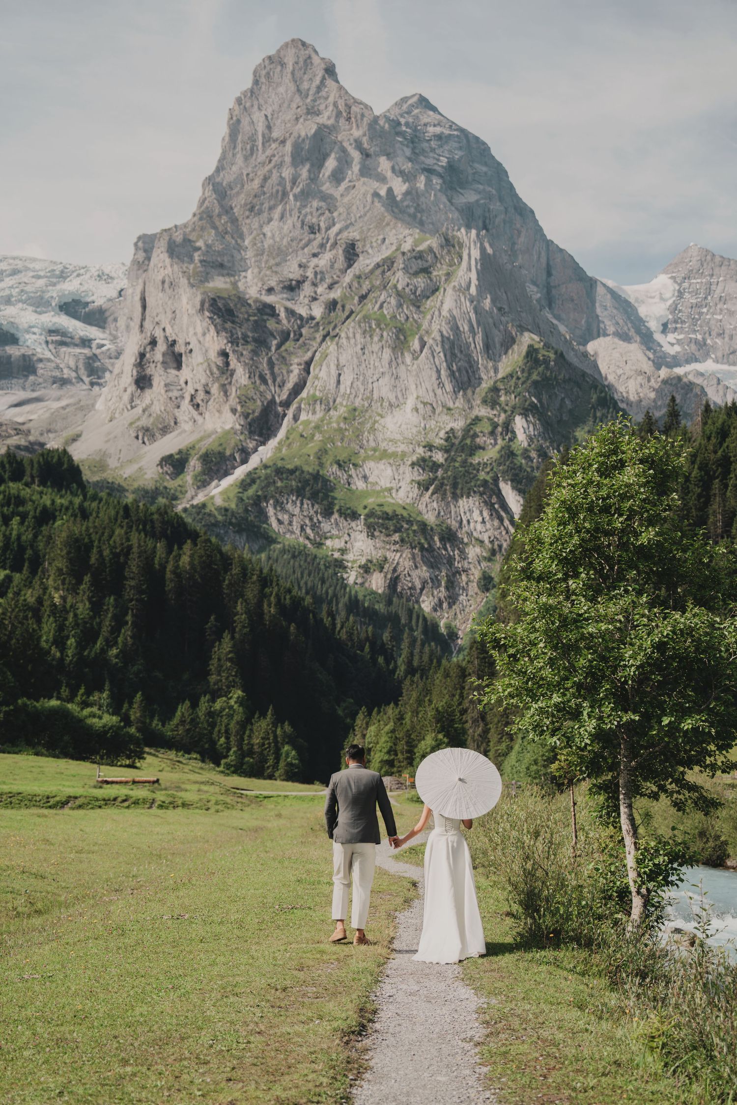 Bride and groom walking toward dramatic Alpine peak on a scenic valley path with white parasol.