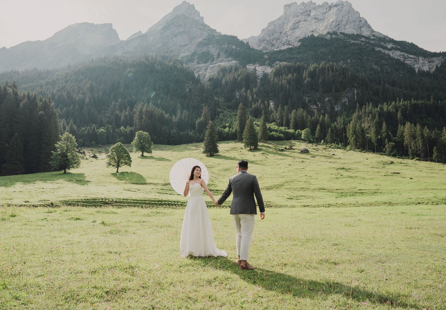 Newlyweds walk hand in hand through scenic alpine meadow, bride carrying white parasol with mountain vista behind.
