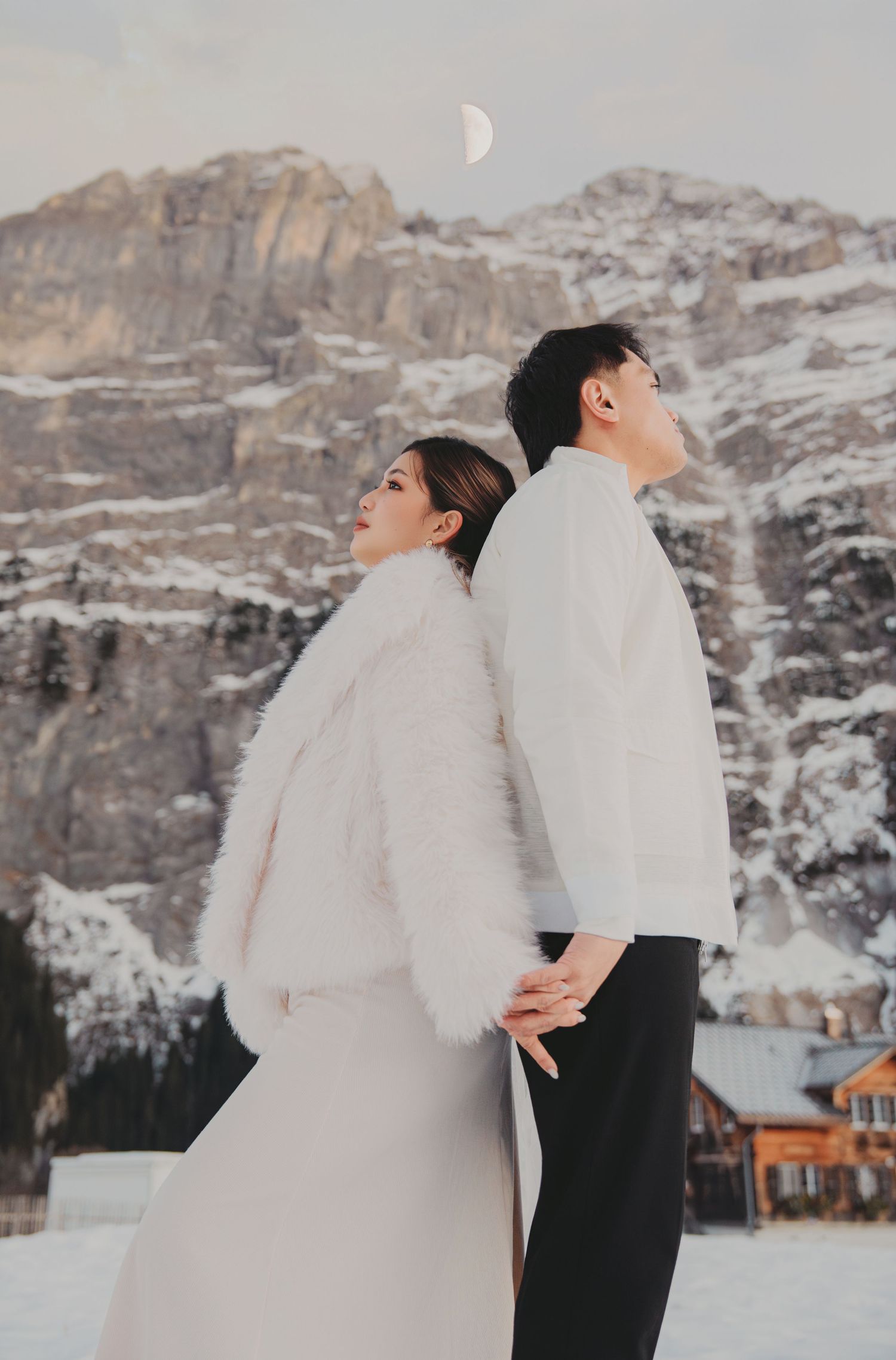 Couple in white winter outfits standing back-to-back holding hands with snowy Dolomites mountain backdrop.