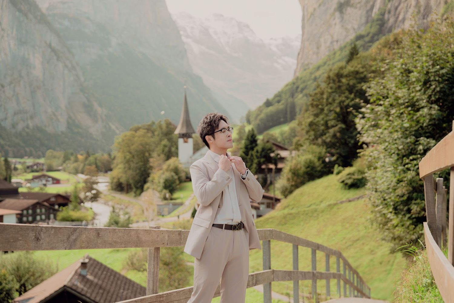Young man in light suit adjusting collar on bridge overlooking scenic Swiss mountain valley village.
