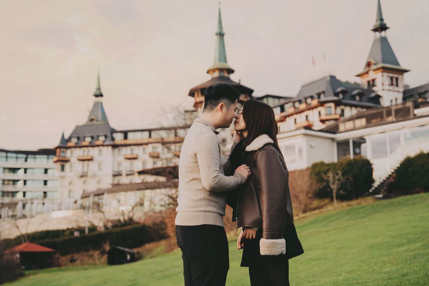 Romantic Asian couple standing close together on lawn outside grand hotel.