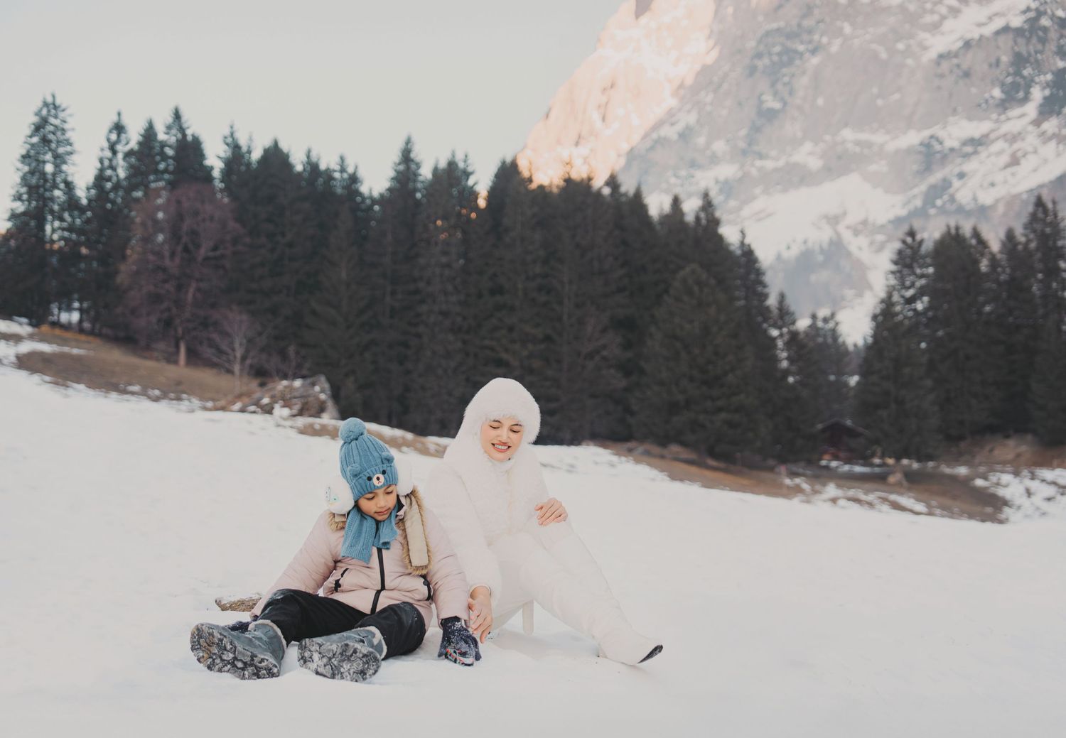 Mother and young child sitting together in snow with pine trees and sunlit mountain peak behind them.