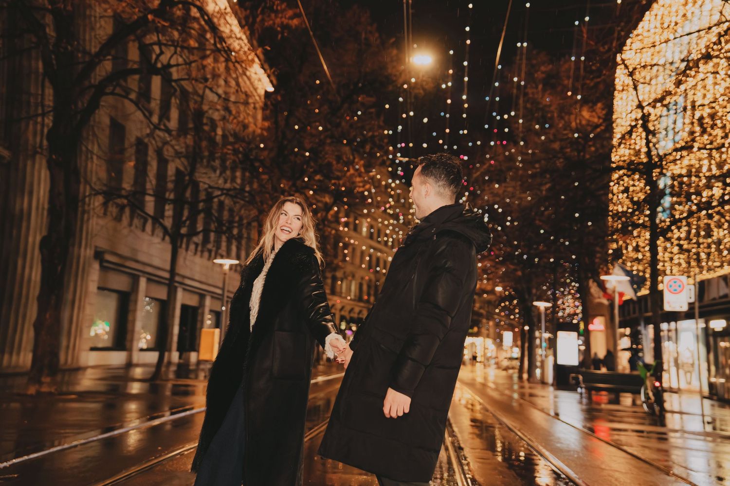 Romantic couple walking hand-in-hand under a canopy of golden Christmas lights on a wet city street.