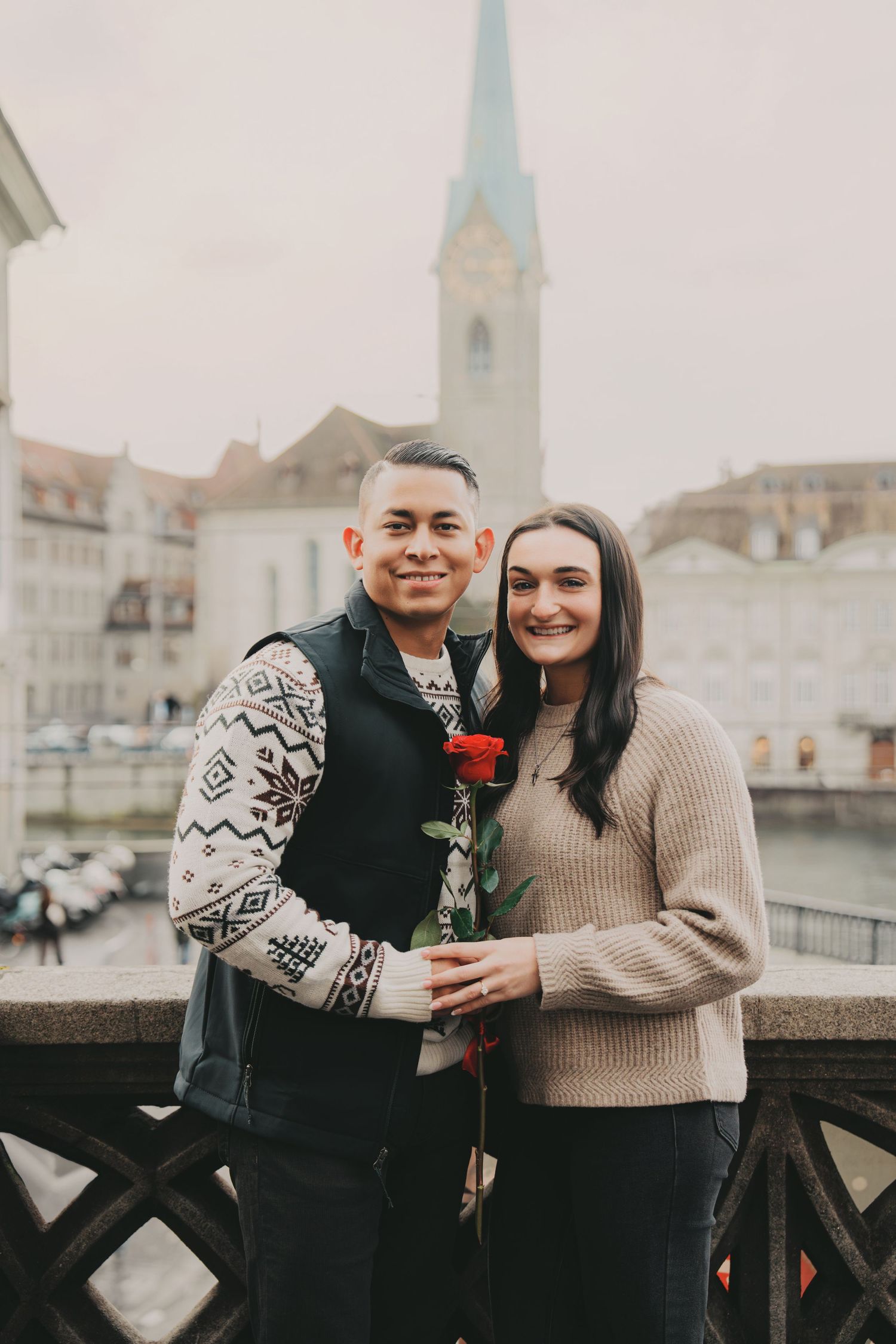 Happy couple posing with a red rose on a bridge with Zurich cityscape behind them.