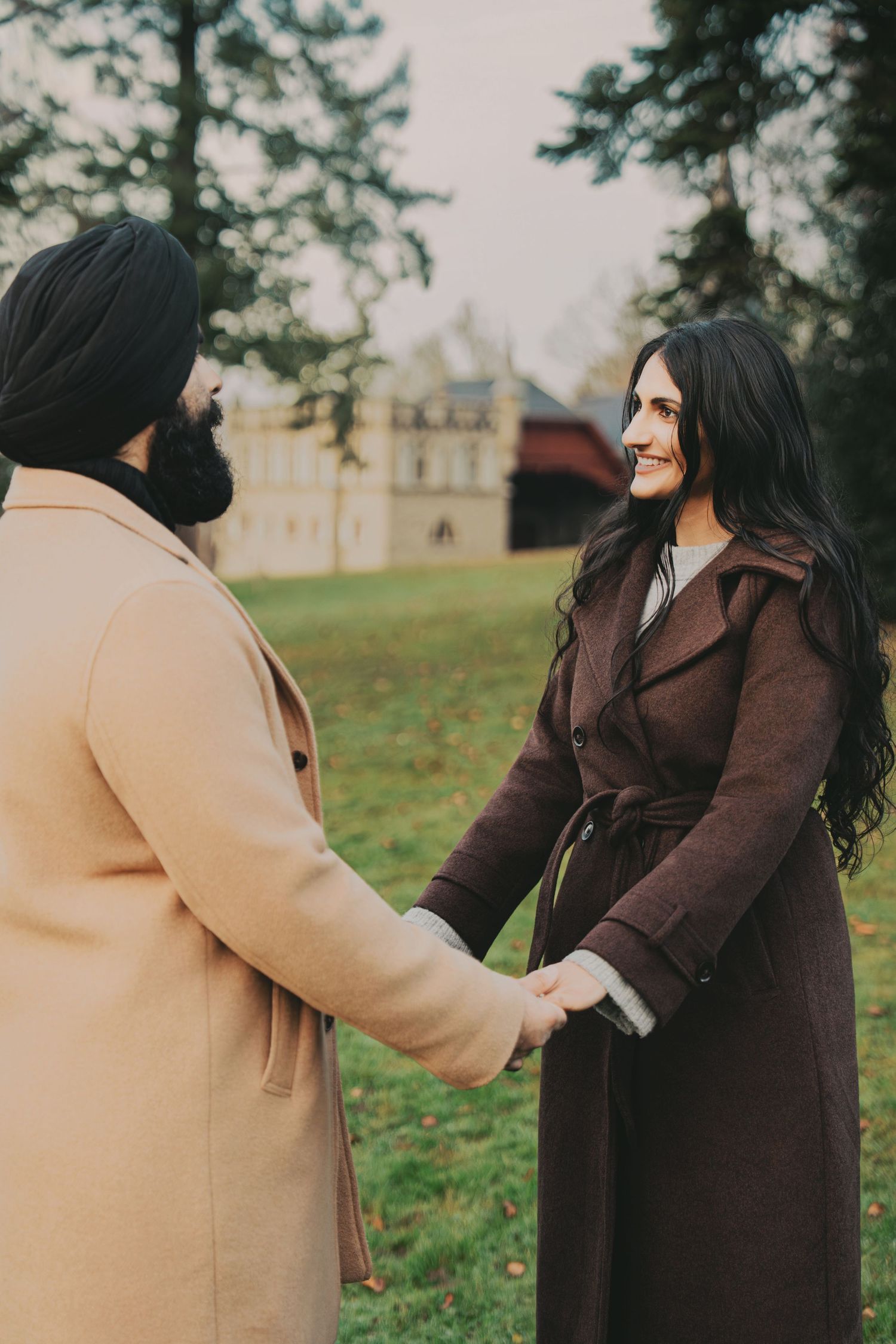 Smiling woman in brown coat holds hands with bearded man outdoors.