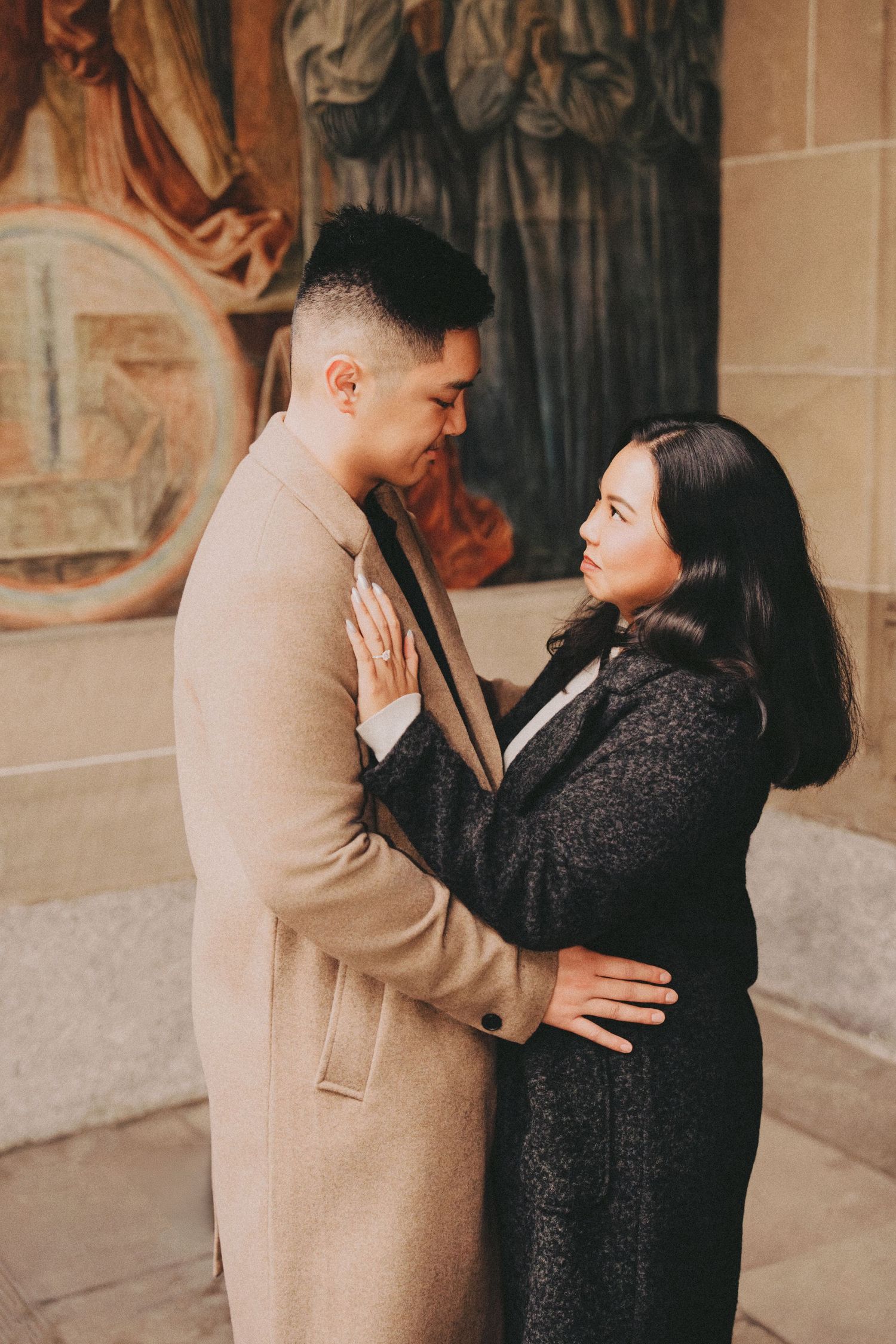 Engaged couple embracing tenderly in front of colorful religious frescos on a stone wall.