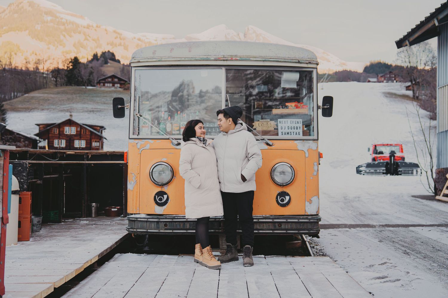 Romantic couple embracing in front of vintage orange food truck in snowy Swiss mountain village.