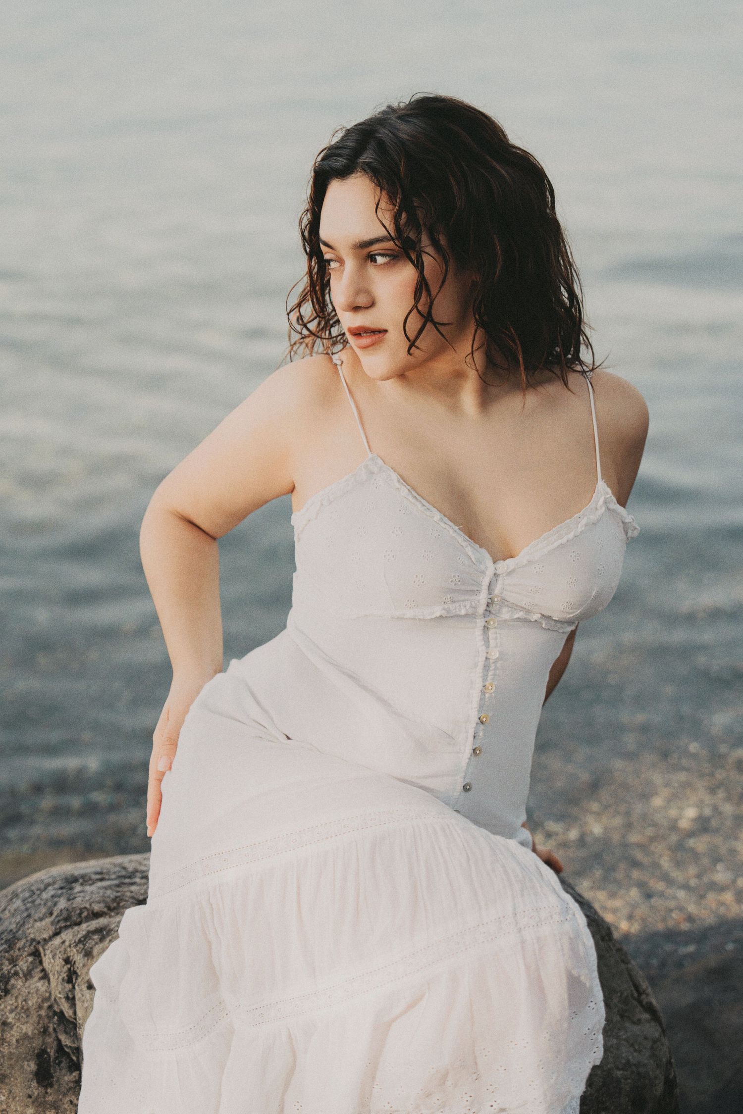 Young woman in white sundress sitting on rocks near the sea, looking away.