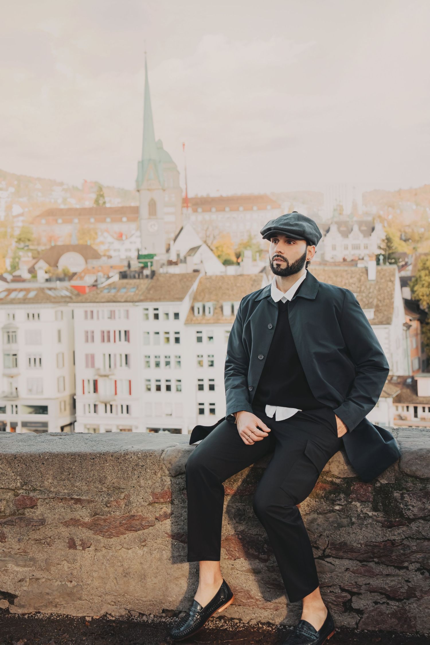 Fashionable bearded man in dark coat and flat cap posing on a stone wall with Zurich skyline behind.