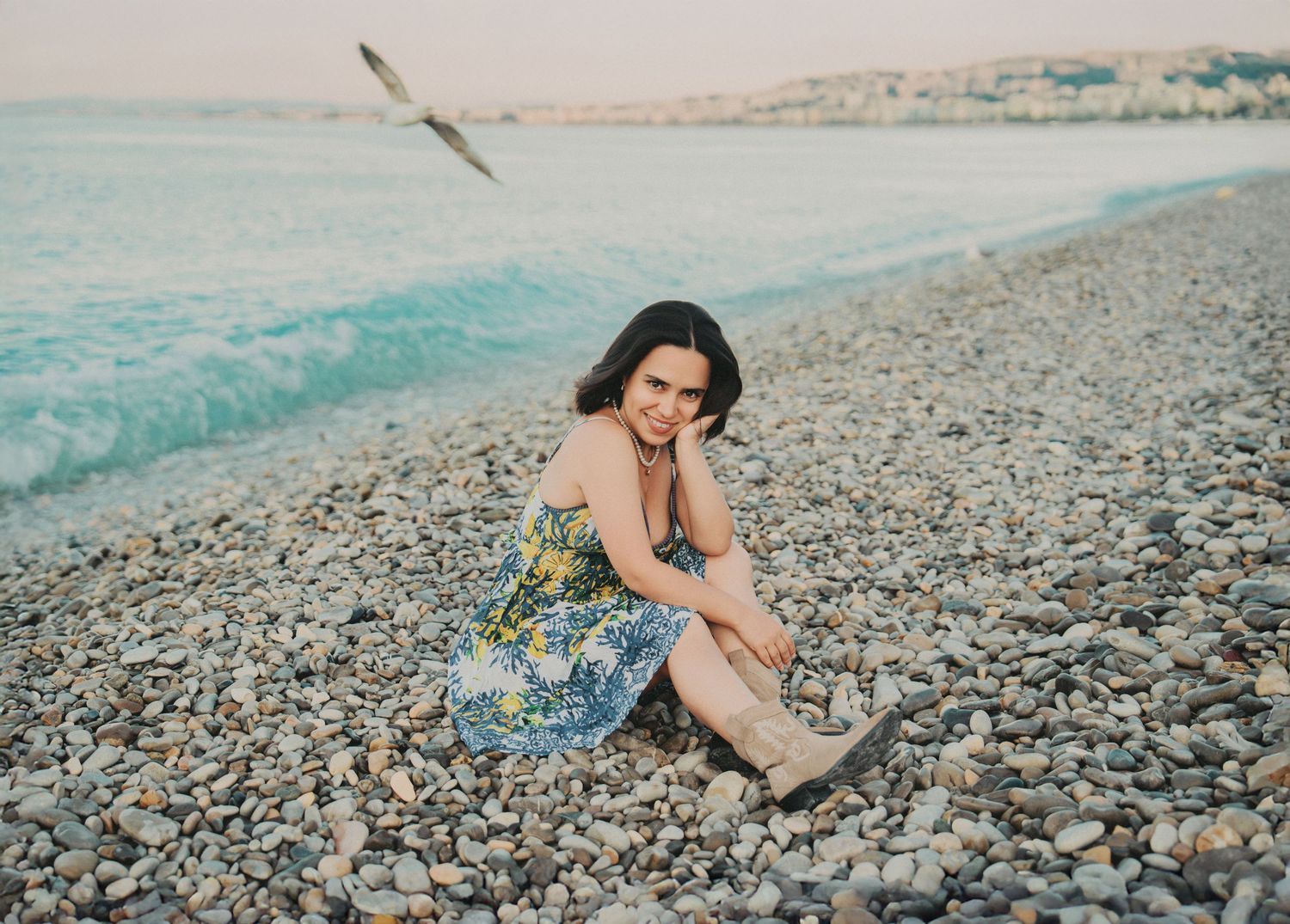A woman in a floral dress sits pensively on a pebble beach as a seagull flies by.