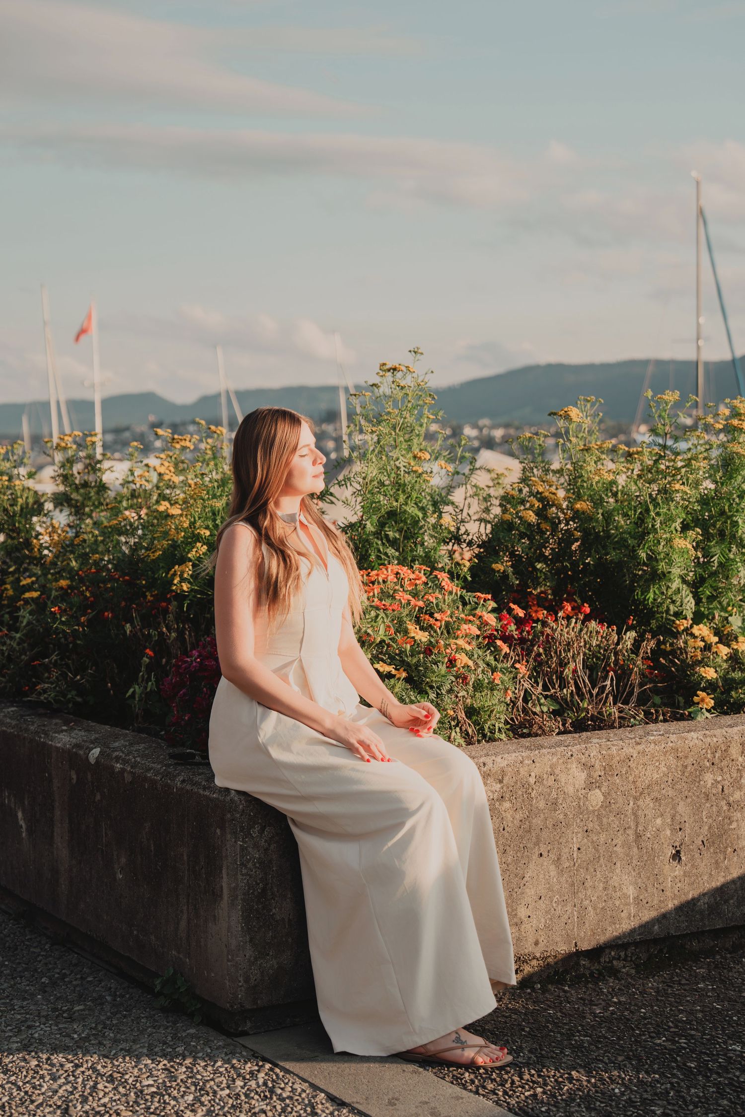 Woman in white dress sitting on stone wall surrounded by colorful flowers near lake.