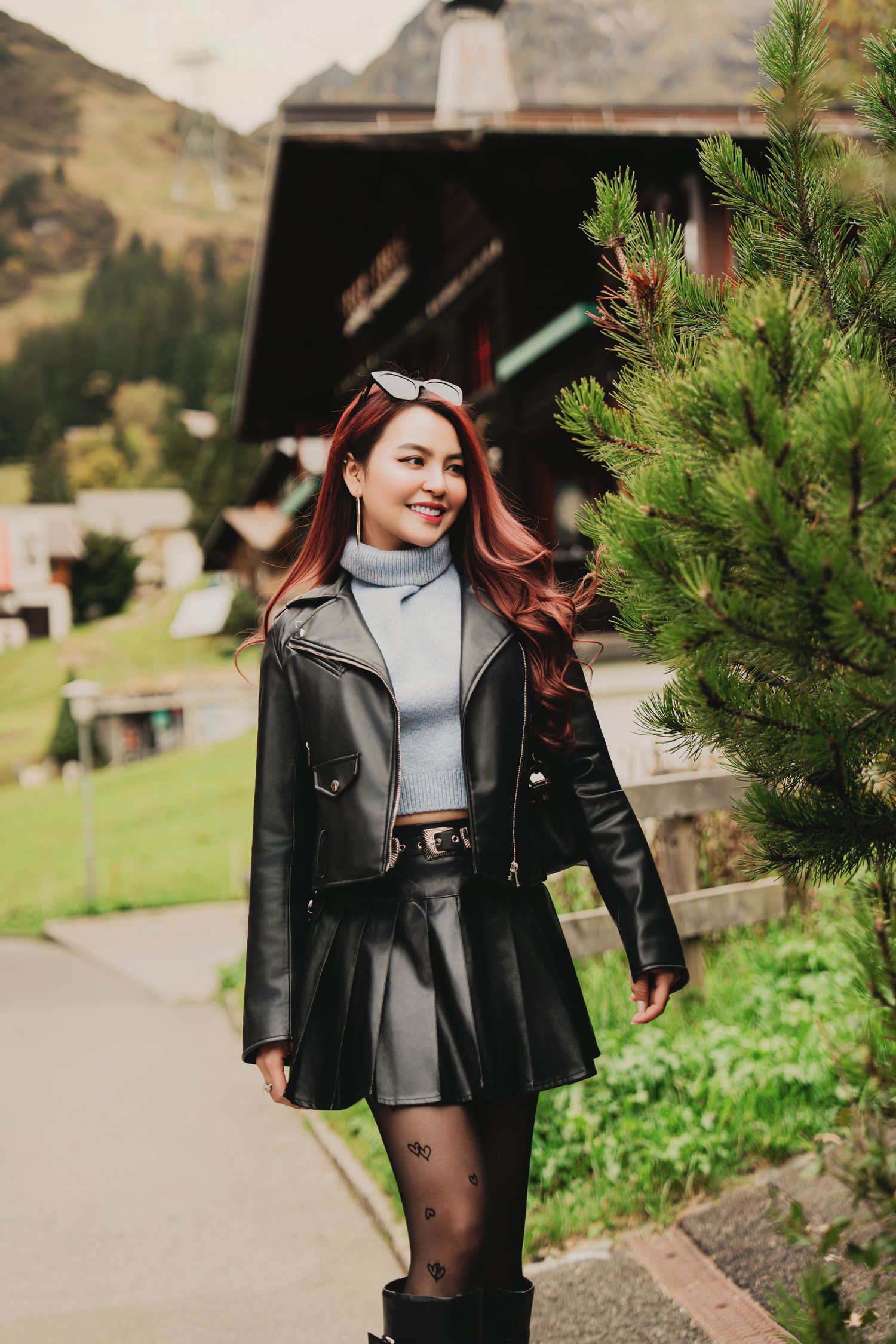 Happy woman with red hair in black leather jacket posing beside a pine tree in an alpine village.