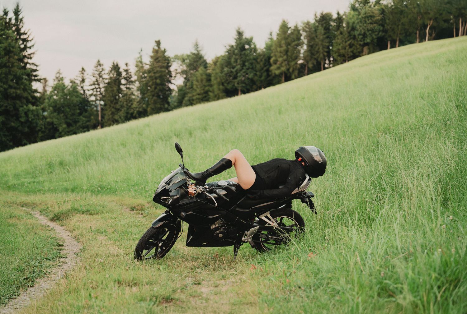 Person lying across black sport motorcycle on grassy hillside path with trees in background.