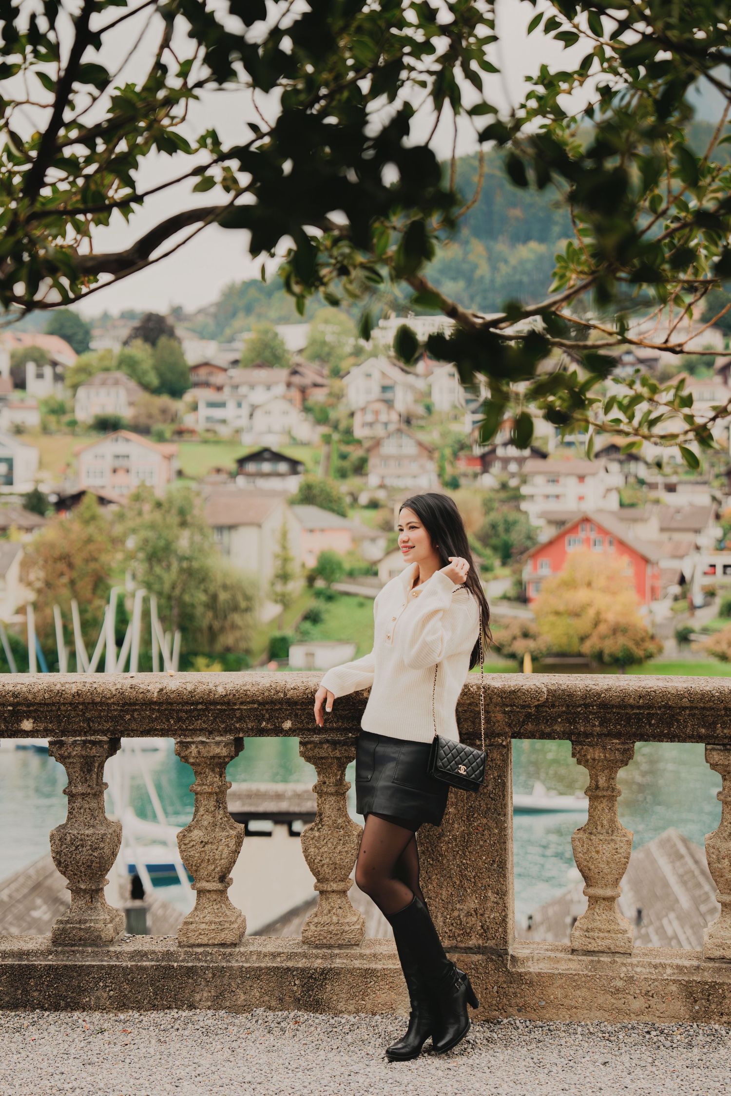 Stylish woman with Chanel bag leans on stone railing with picturesque Swiss lakeside village behind.