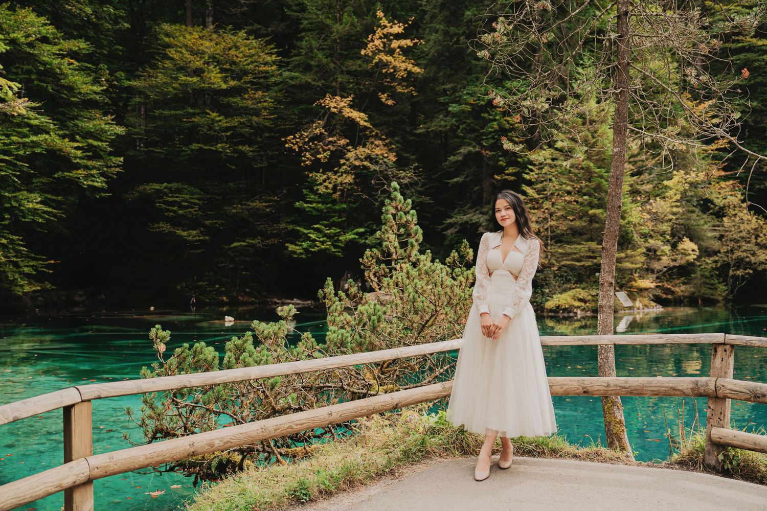 Elegant woman in white tulle dress holding flowers beside a wooden fence near a bright turquoise lake.