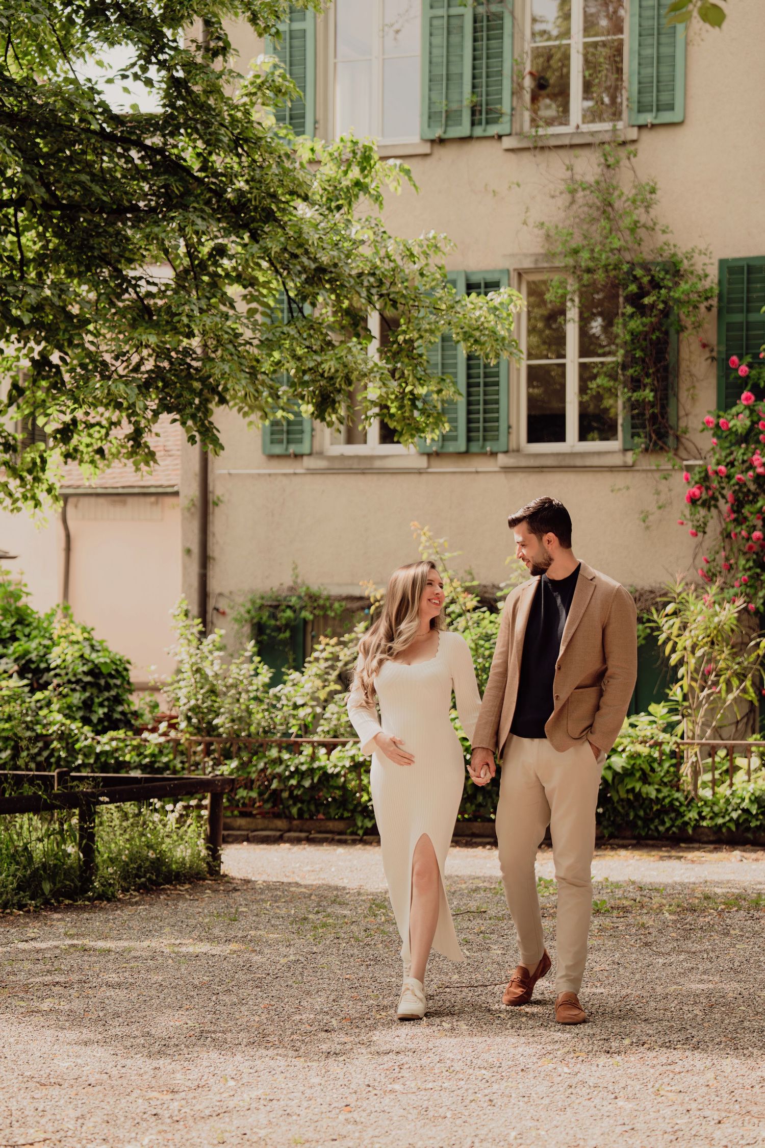Pregnant couple holding hands walking through a lush garden path beside a charming European building.