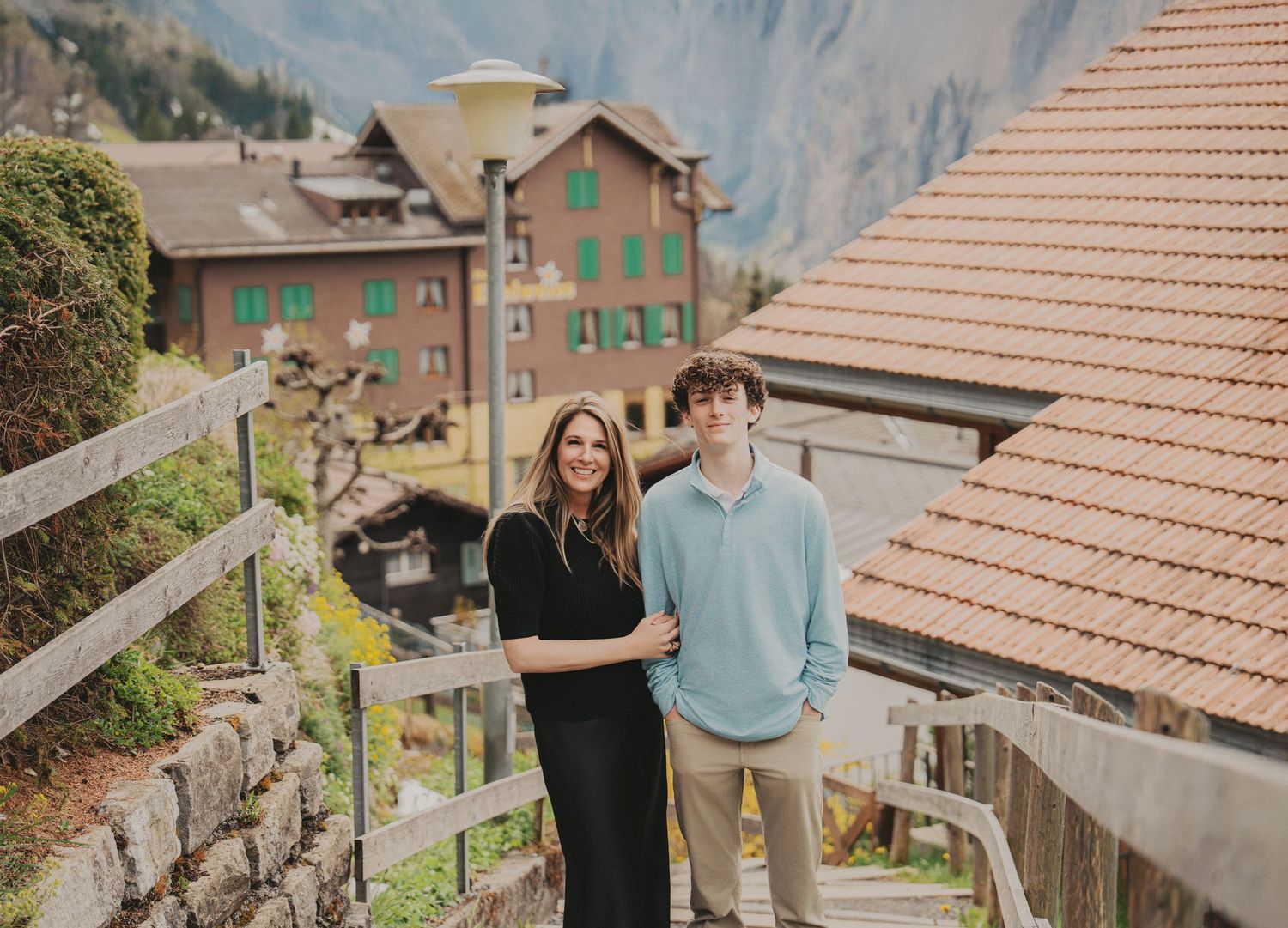 Smiling mother and son standing close together on stairs with Swiss chalet buildings behind them.