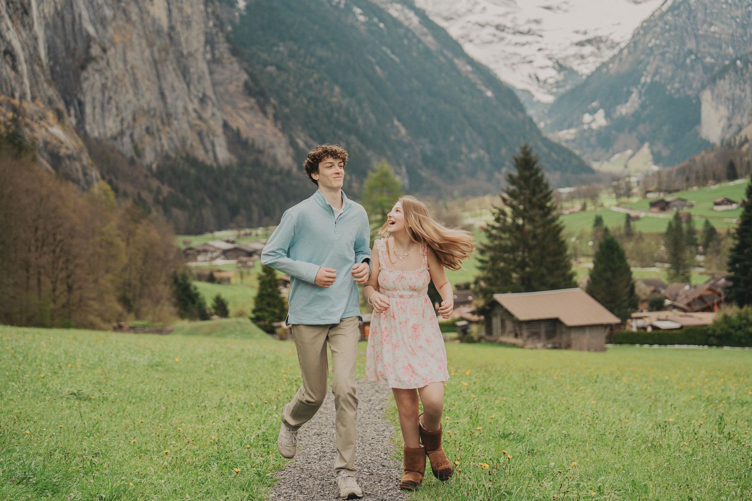 Laughing couple strolling along a gravel path in a beautiful Alpine valley with dramatic mountain cliffs and village scenery.