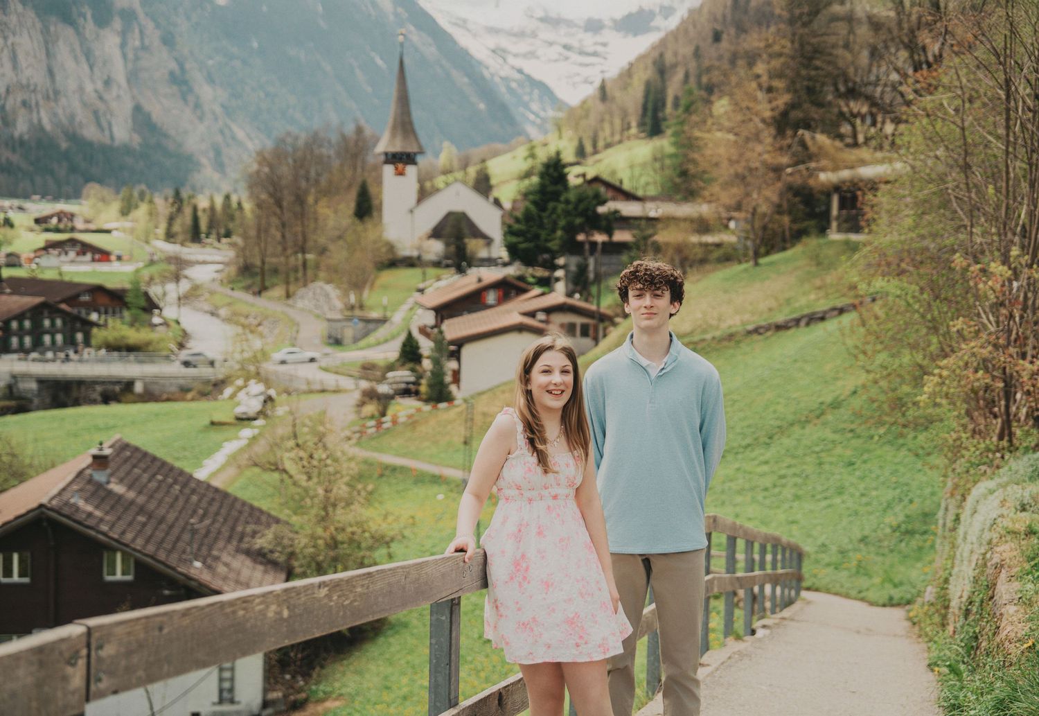 Teen siblings smiling together on a wooden bridge with a scenic Swiss village church and mountains behind them.