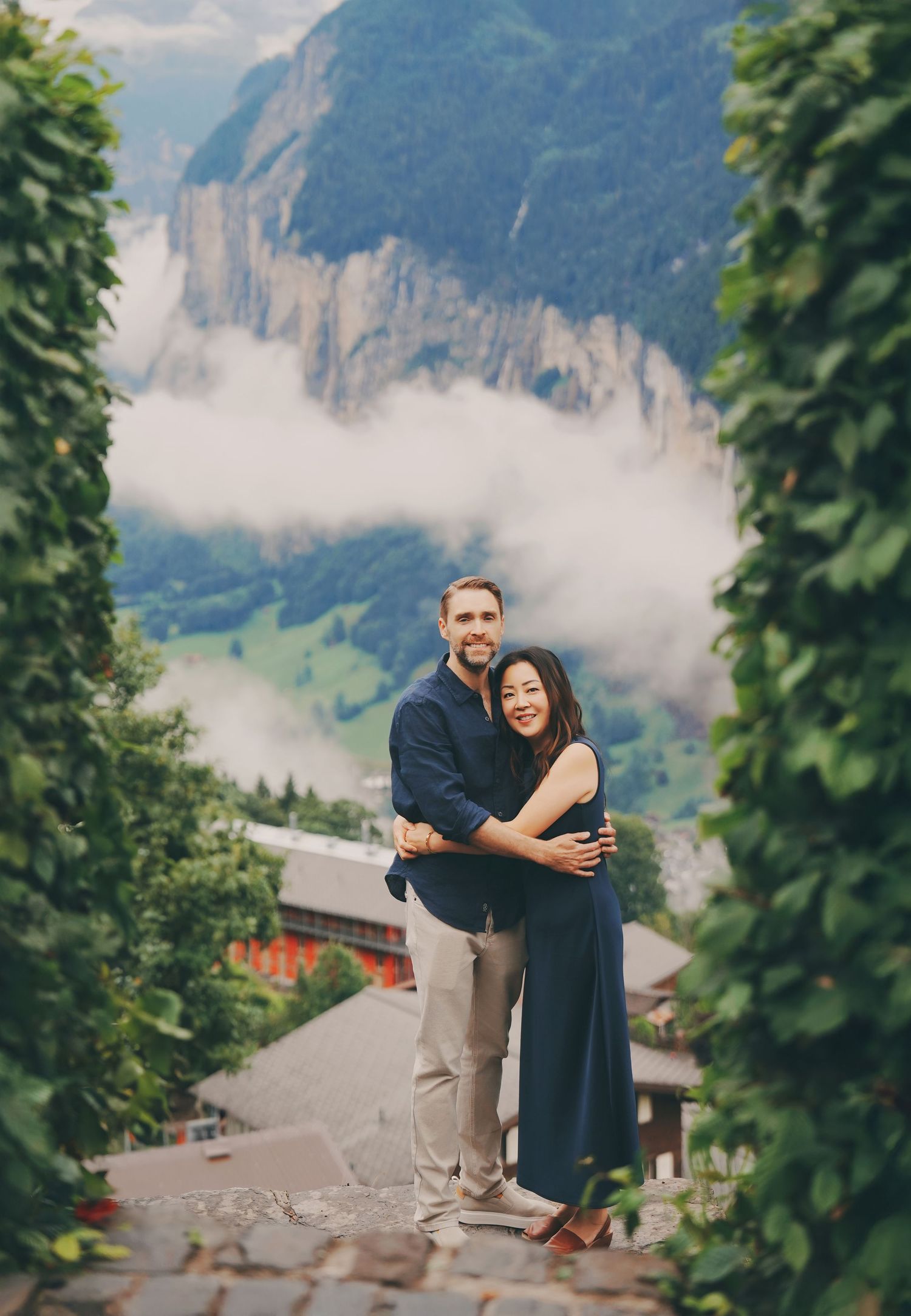 Couple embraces on cobblestone path framed by lush greenery with stunning Swiss Alpine valley view.