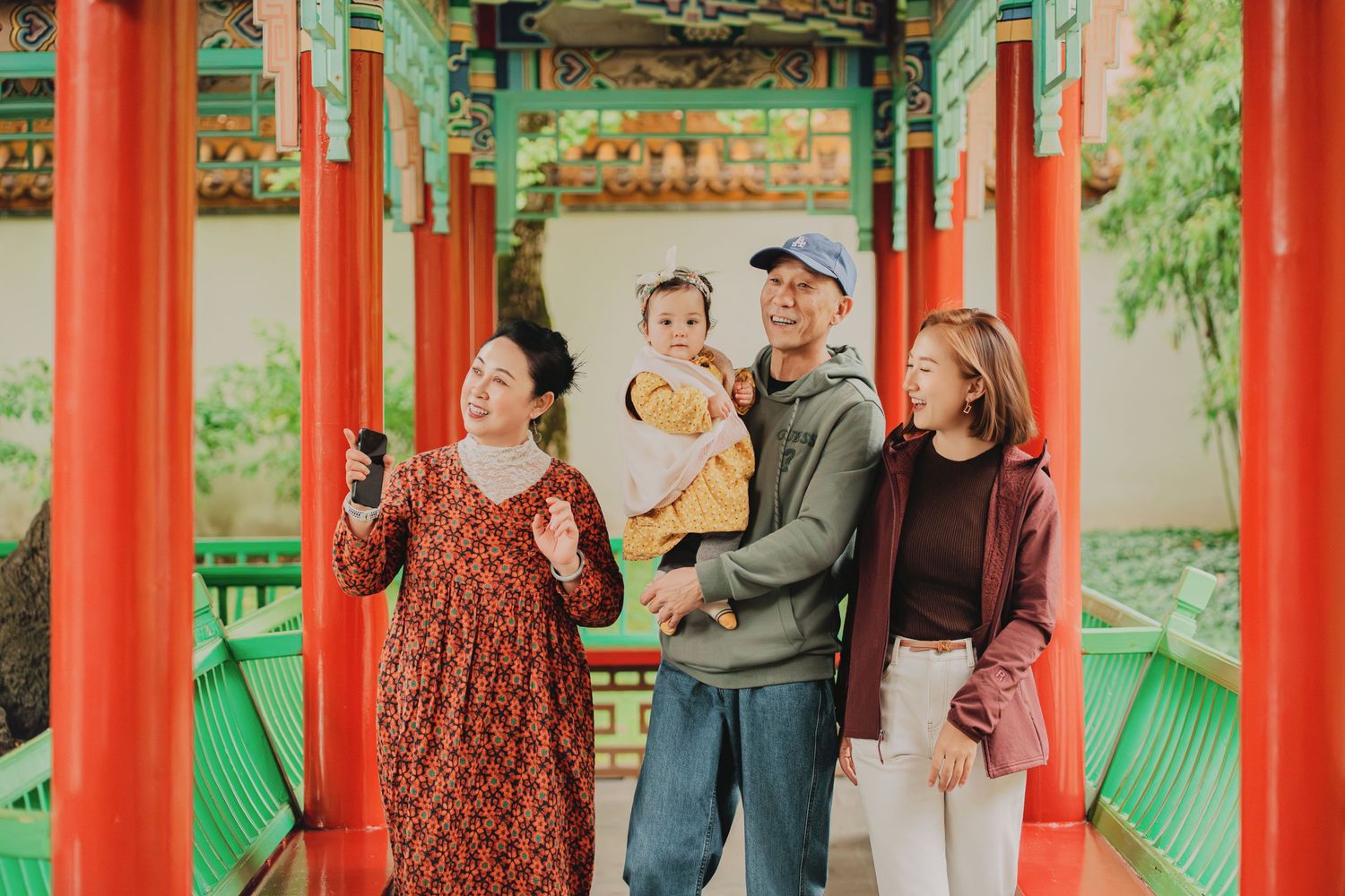 Asian family of four with baby strolling through colorful red Chinese garden corridor together.