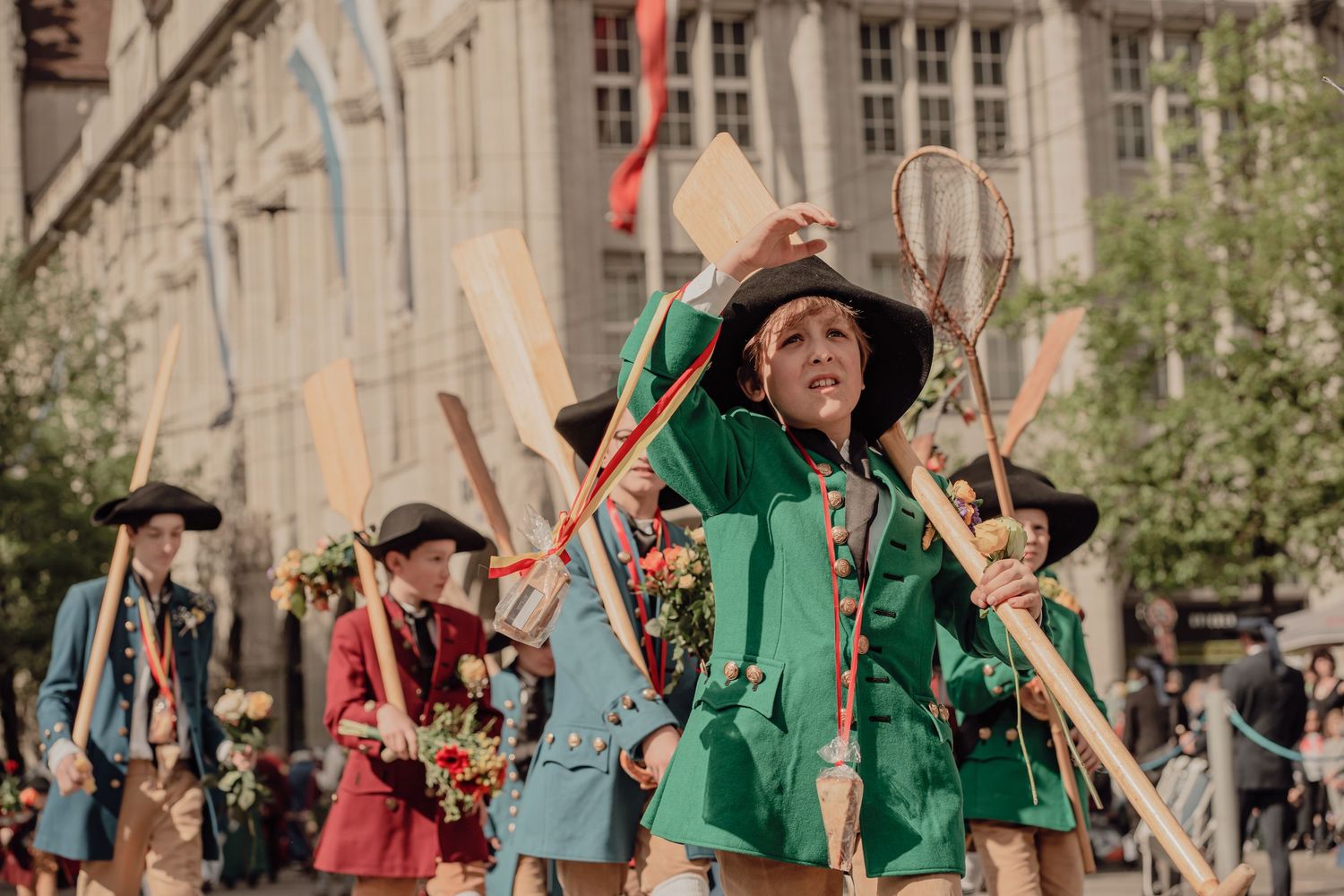 A child in a green medieval costume leads performers carrying long sticks in a colorful historical parade.