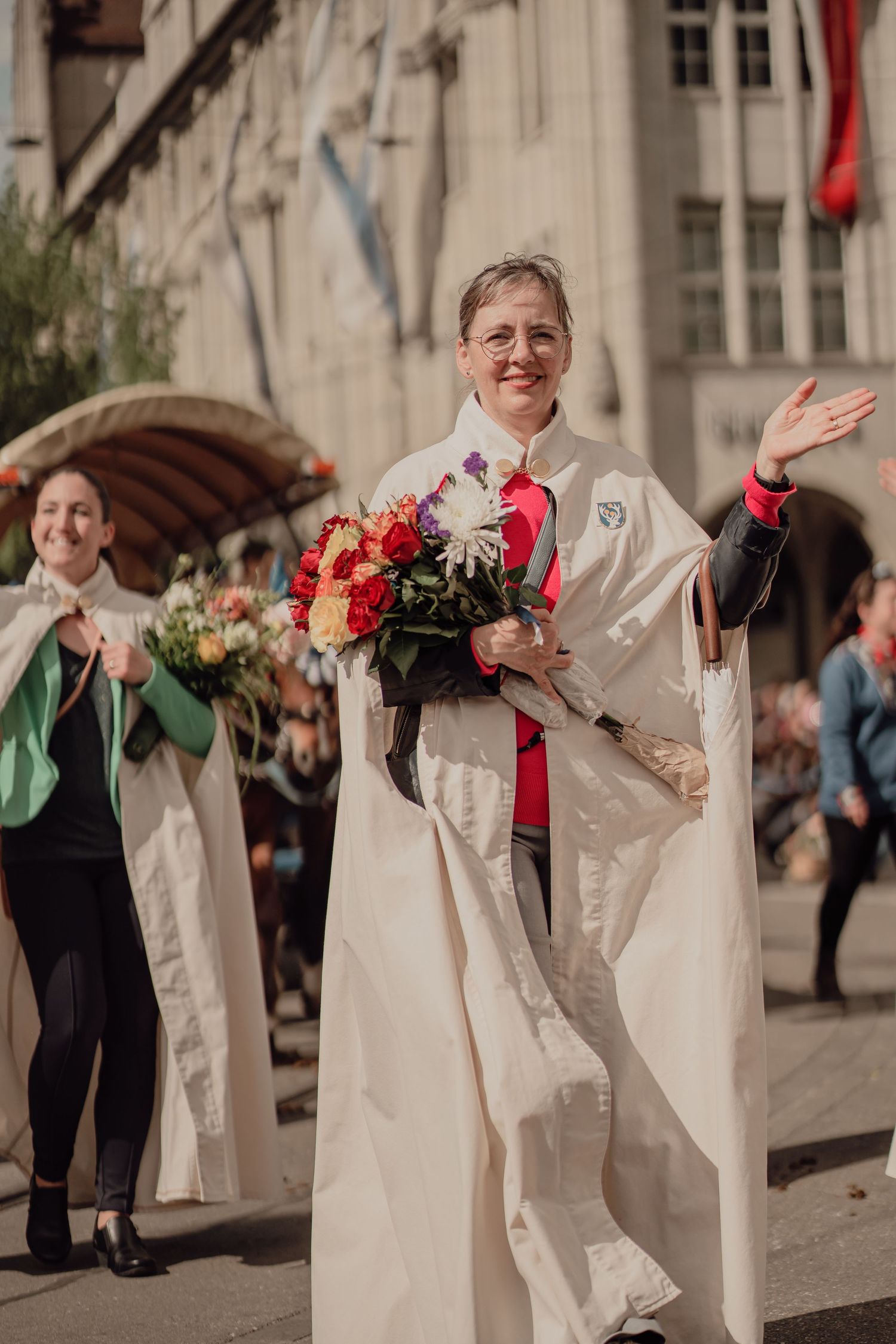 Woman in white ceremonial robe waves holding colorful bouquet during festive street parade.