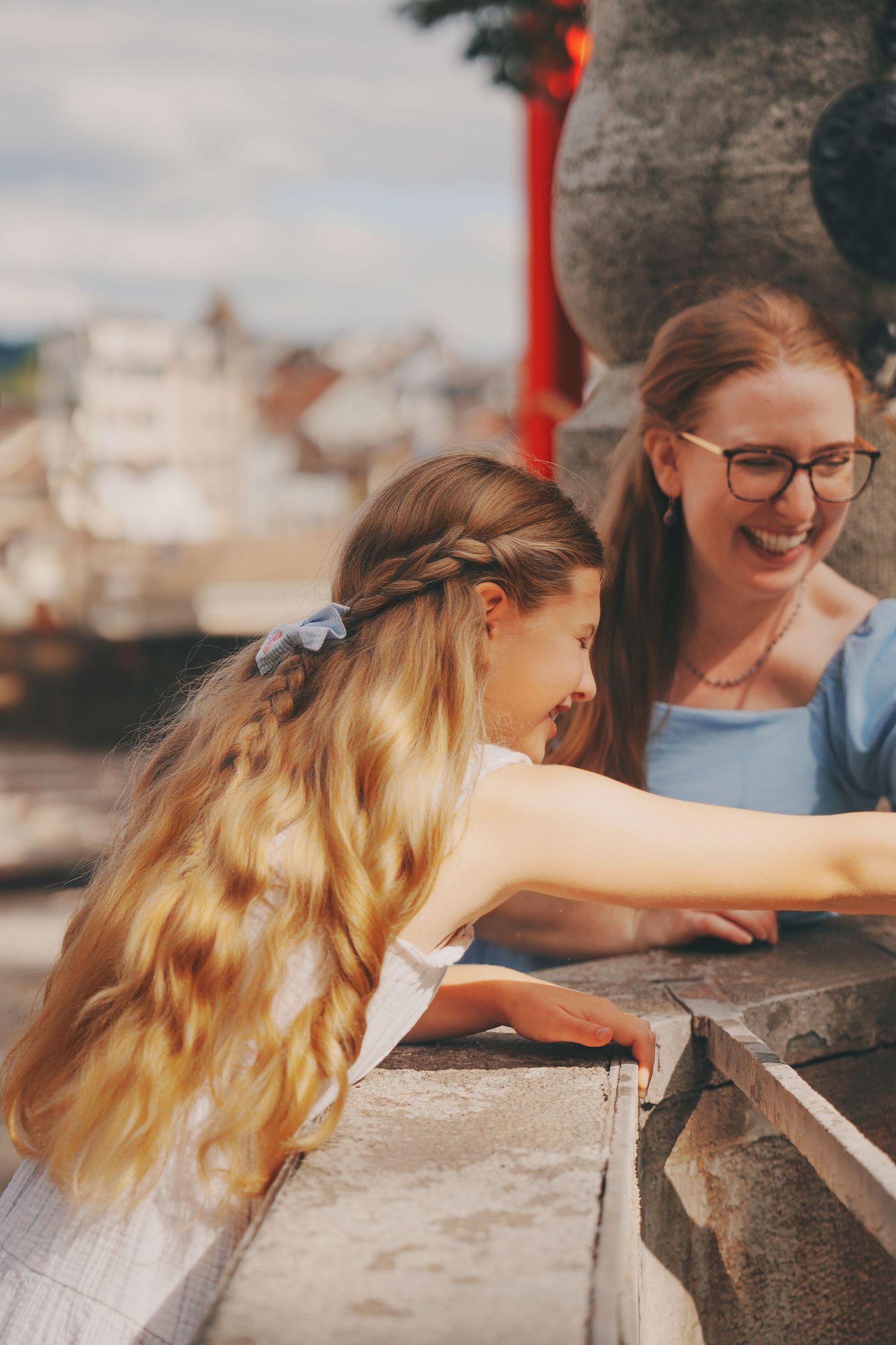 A smiling woman with glasses and a young blonde girl lean over a stone wall outdoors.