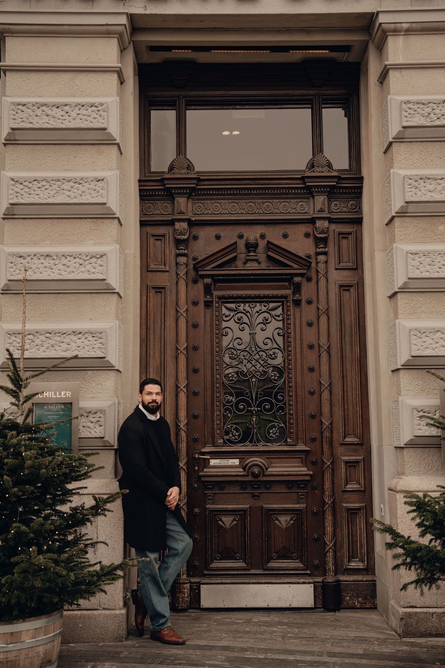 Stylish bearded man standing before grand ornate wooden entrance door of historic European building.