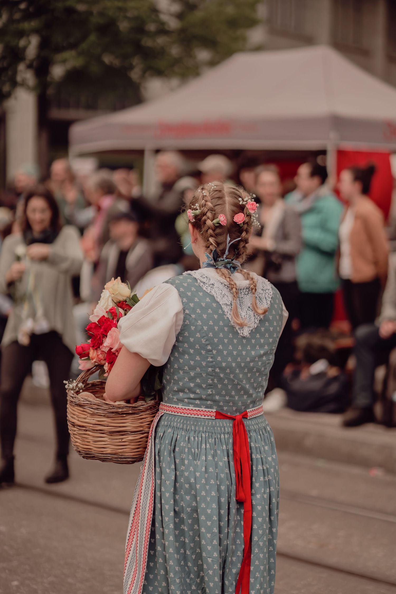 Back view of girl in polka dot dirndl dress carrying wicker basket of flowers at street festival.