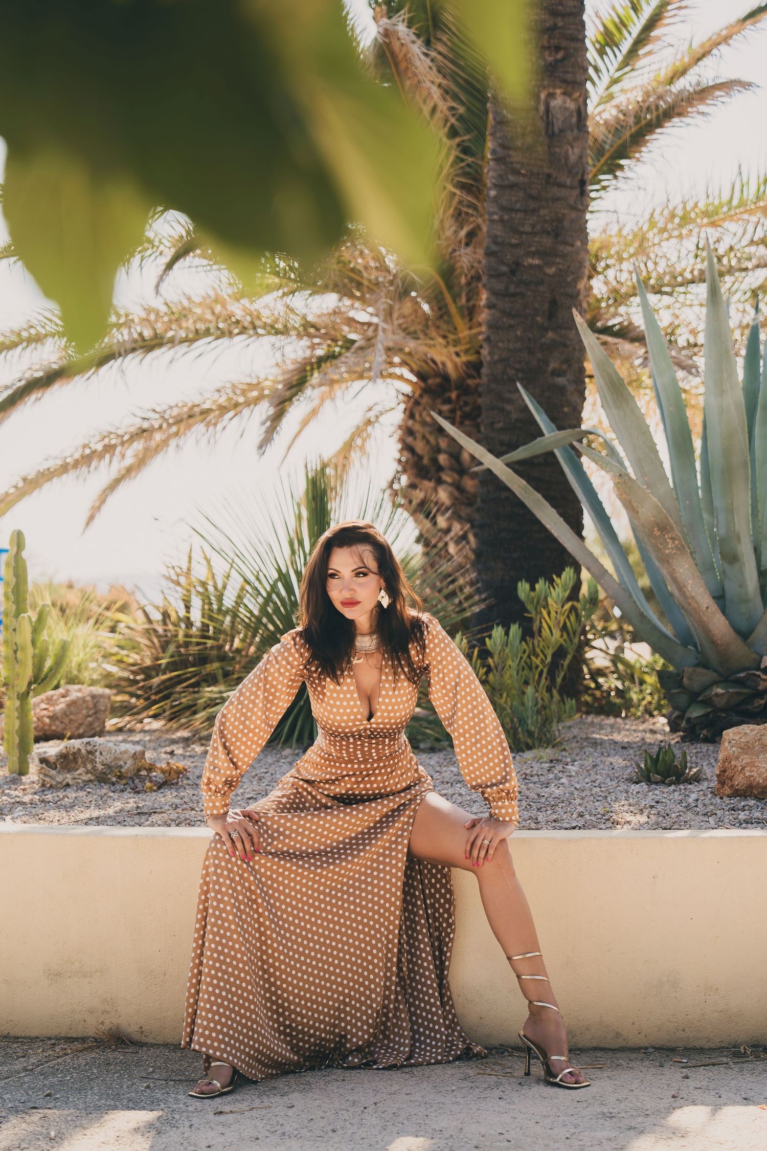 Woman in gold polka dot maxi dress sitting on low wall with palm trees and succulents behind.