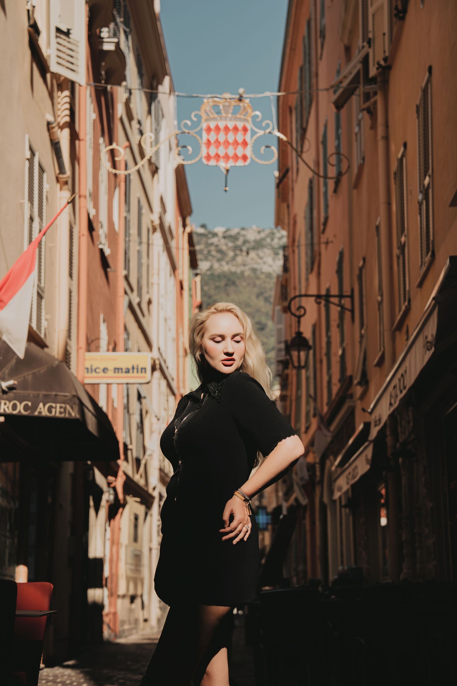Blonde woman in black dress posing in a sunlit European alley with Monaco emblem overhead.
