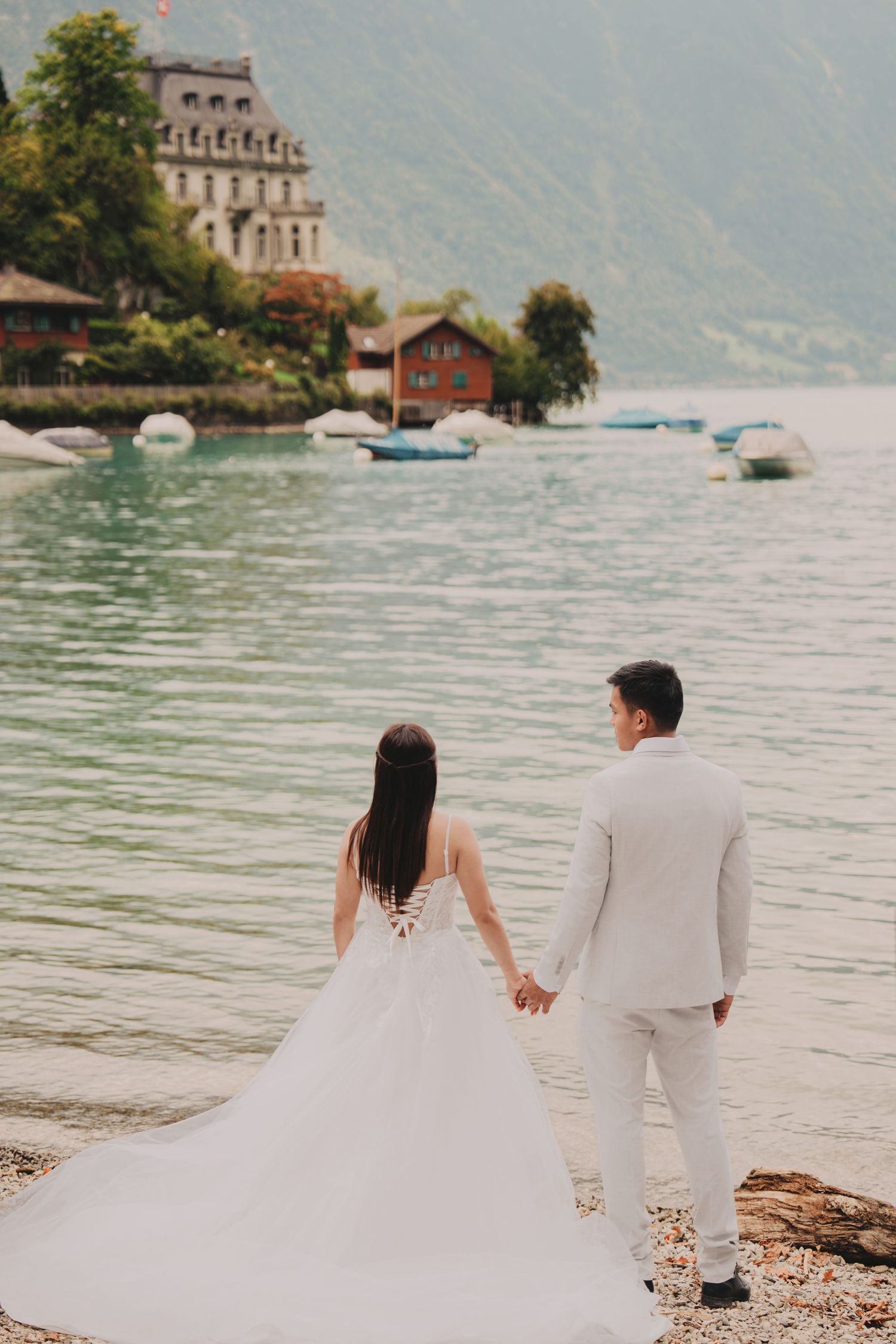 Bride and groom walking hand in hand along a pebbled alpine lakeside with a grand historic building nearby.