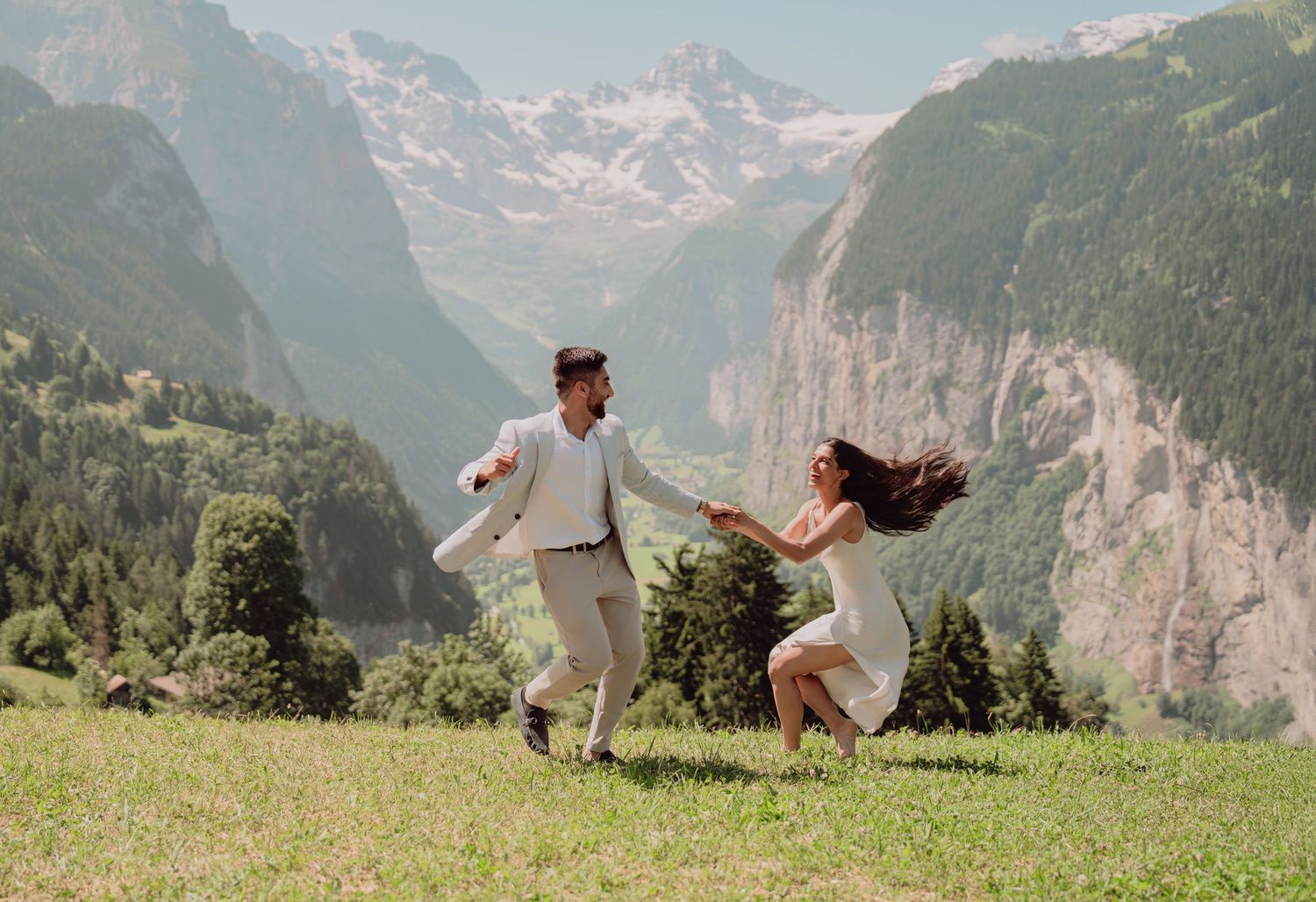 Joyful couple running hand-in-hand on a green alpine meadow with stunning Swiss mountain valley backdrop.