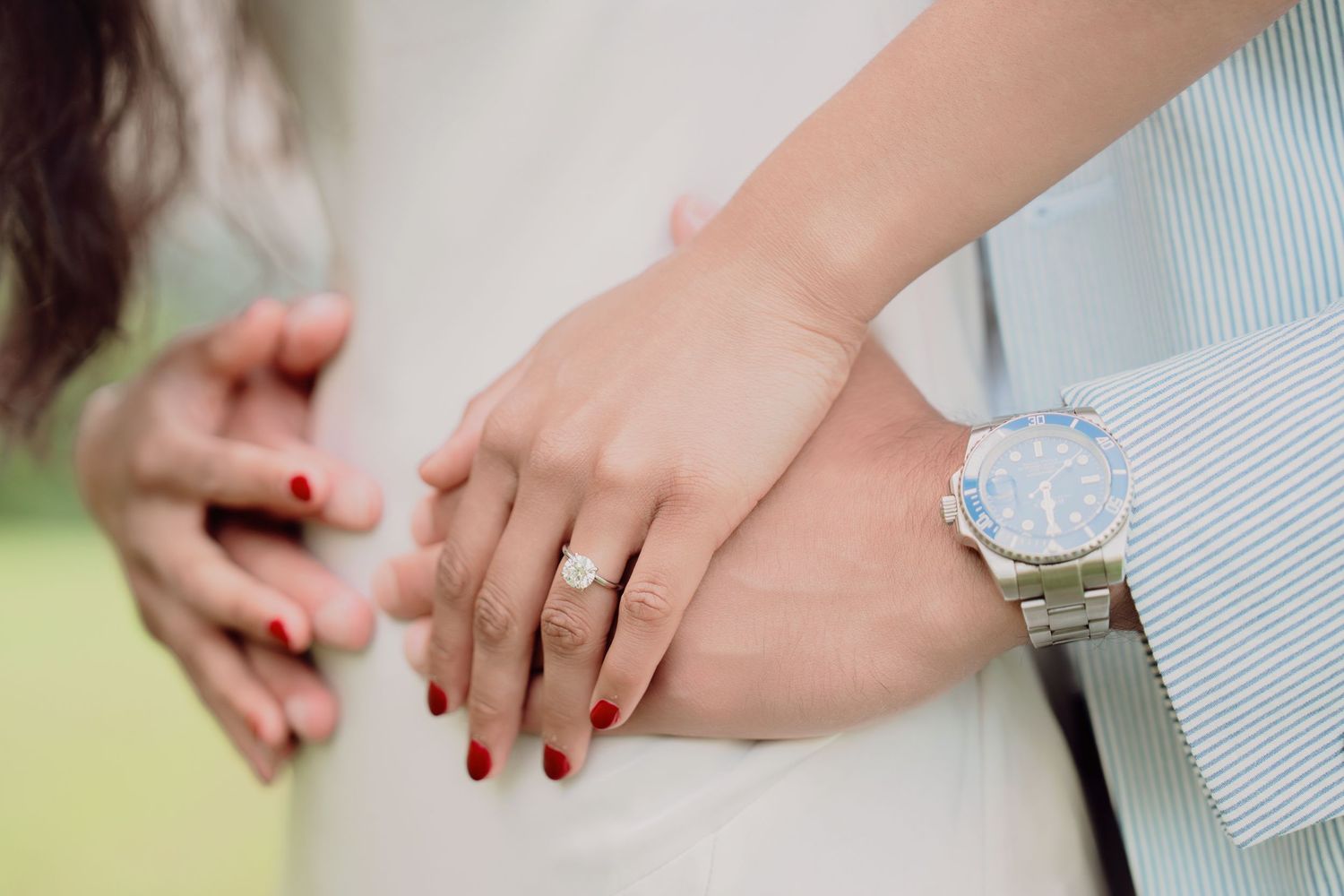 Close-up of couple's hands showing a diamond engagement ring and luxury watch against a soft background.