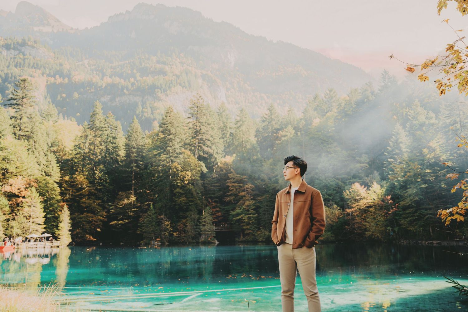 Man with hands in pockets gazing at a glowing turquoise alpine lake in autumn sunshine.