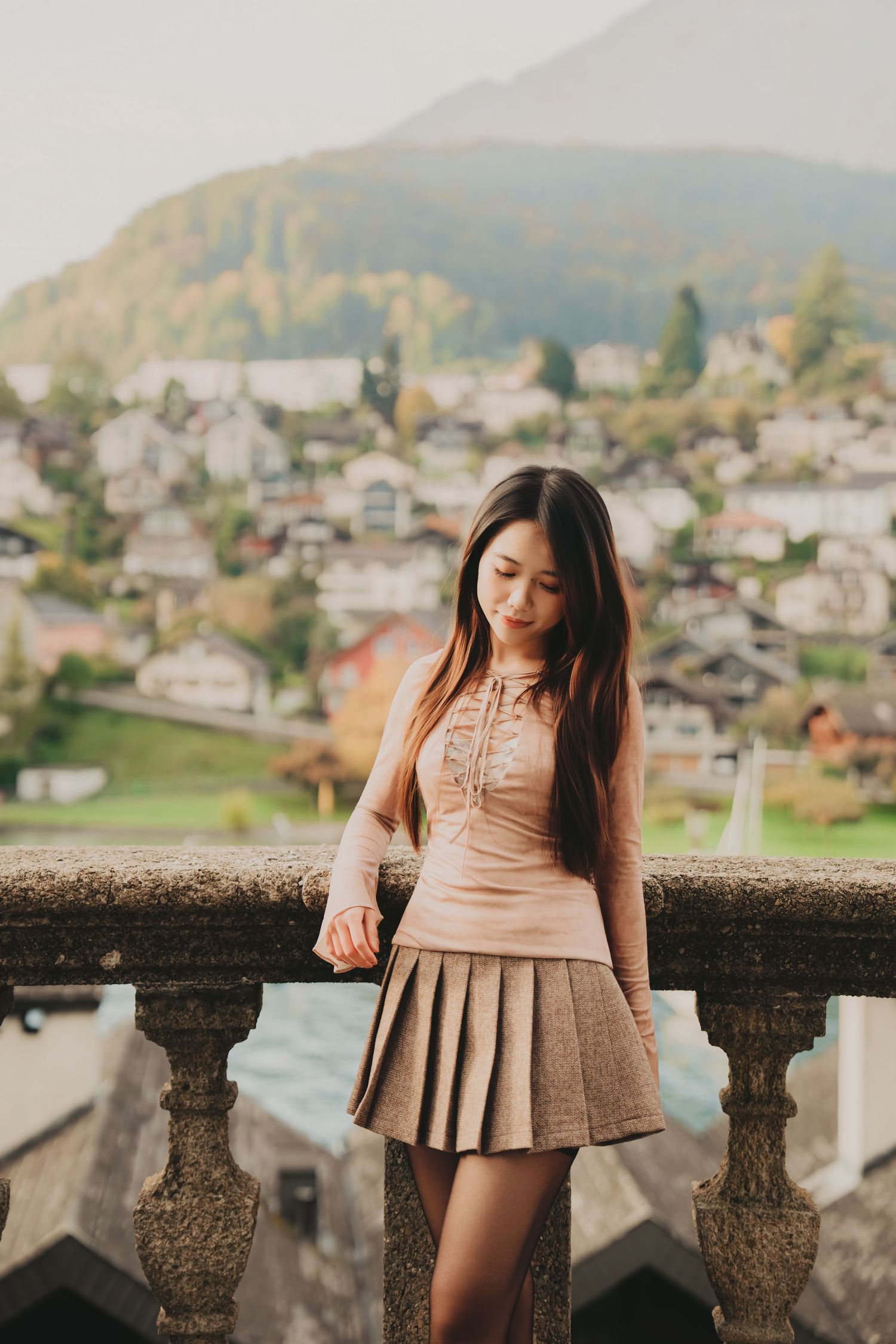 Young Asian woman in beige top and pleated skirt poses on stone balcony with European mountain village backdrop.