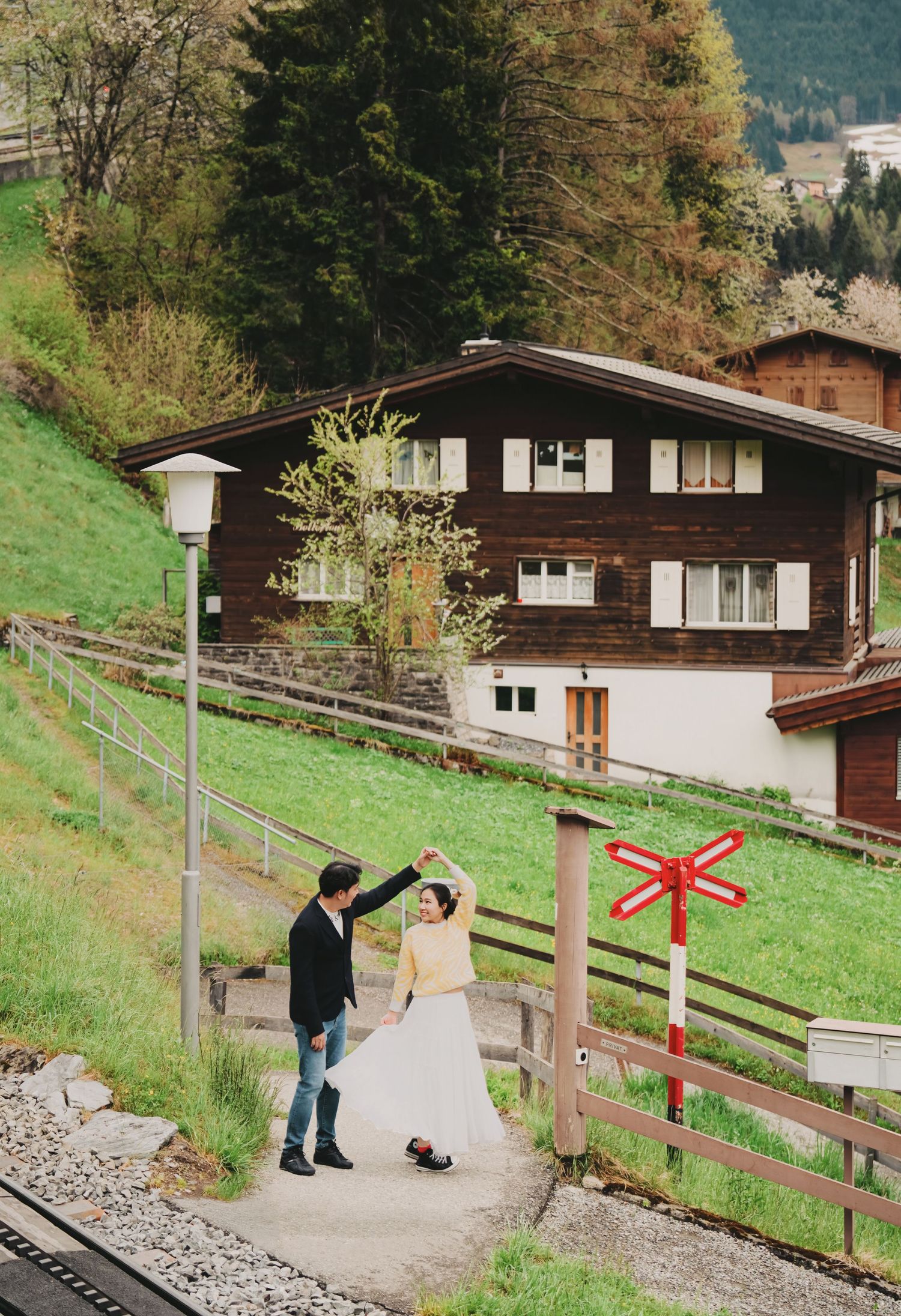 Couple dancing near railway tracks with a traditional Swiss chalet and green hillside in the background.