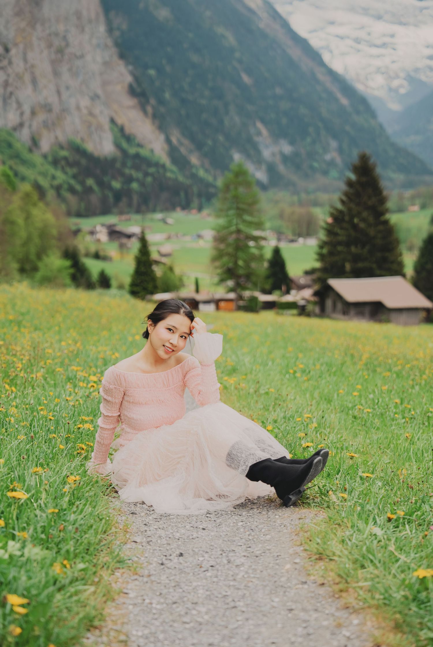 Smiling young woman in pink dress sitting on path surrounded by yellow wildflowers in Swiss alpine valley.